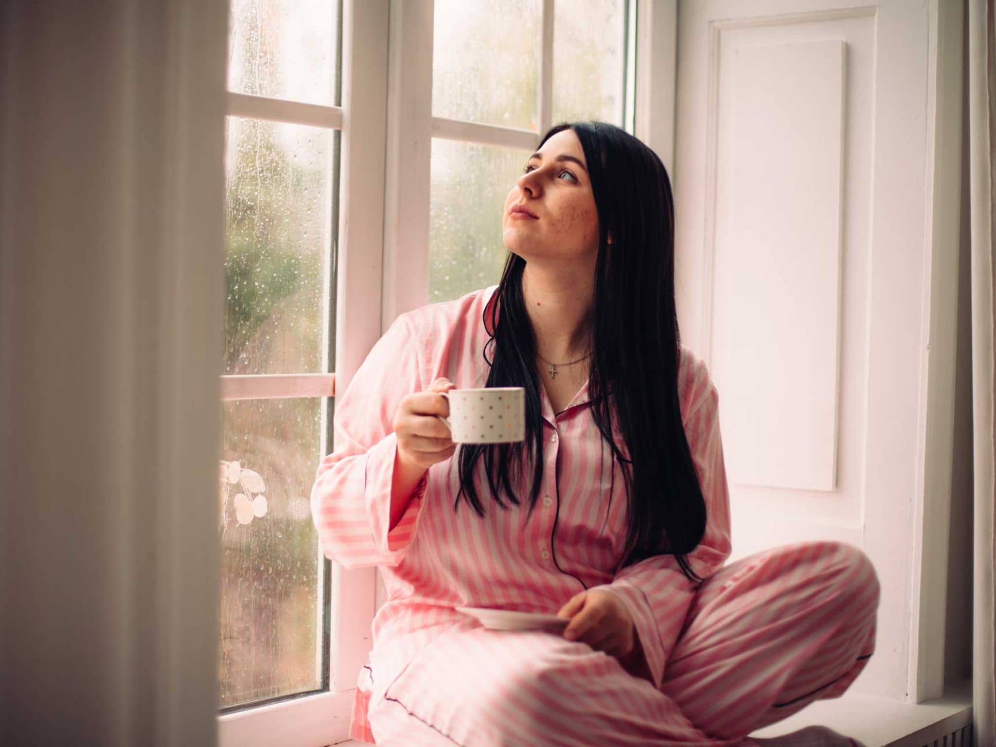 Junge Frau sitzt am Fenster und hält eine Tasse Kaffee