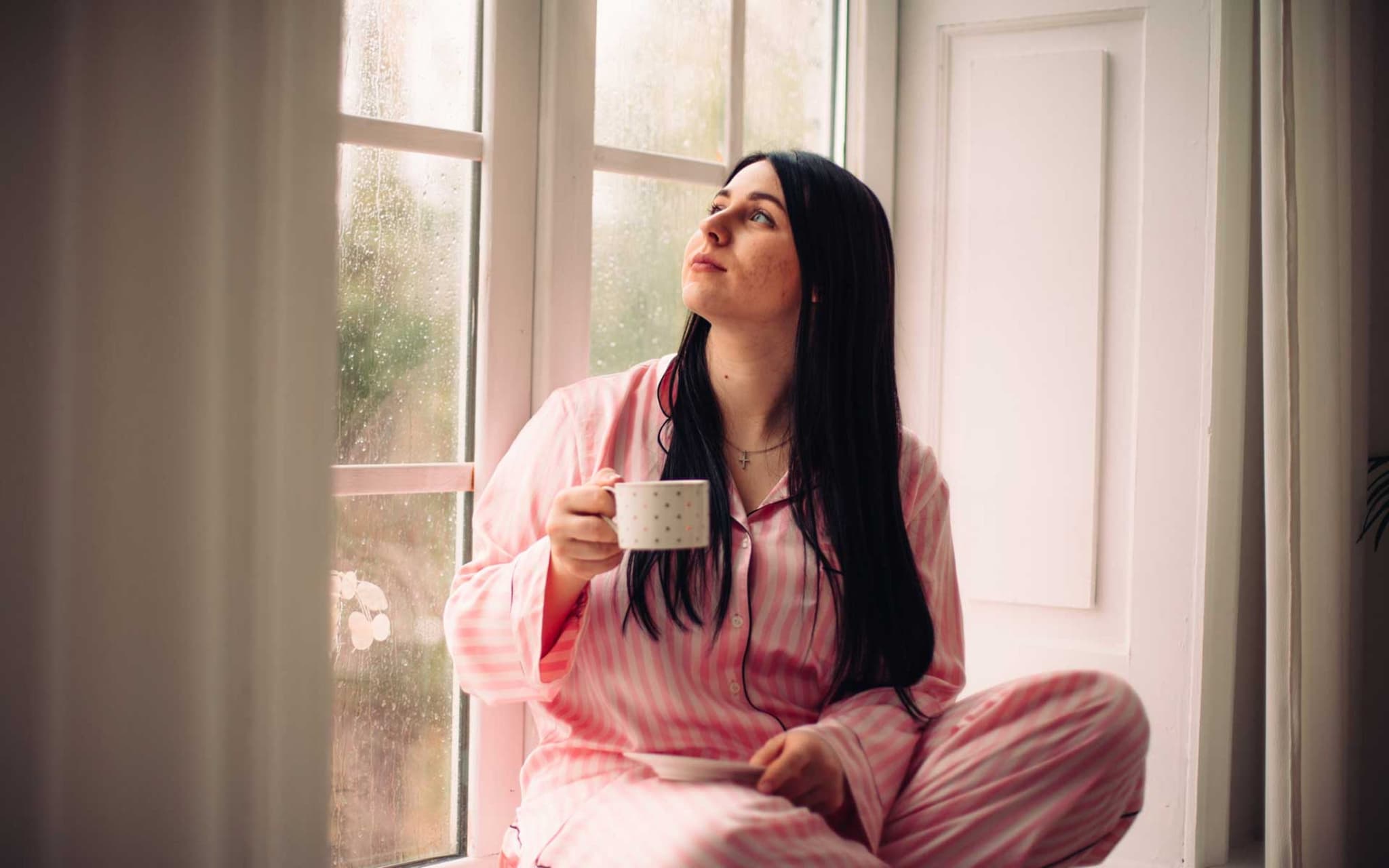 Junge Frau sitzt am Fenster und hält eine Tasse Kaffee