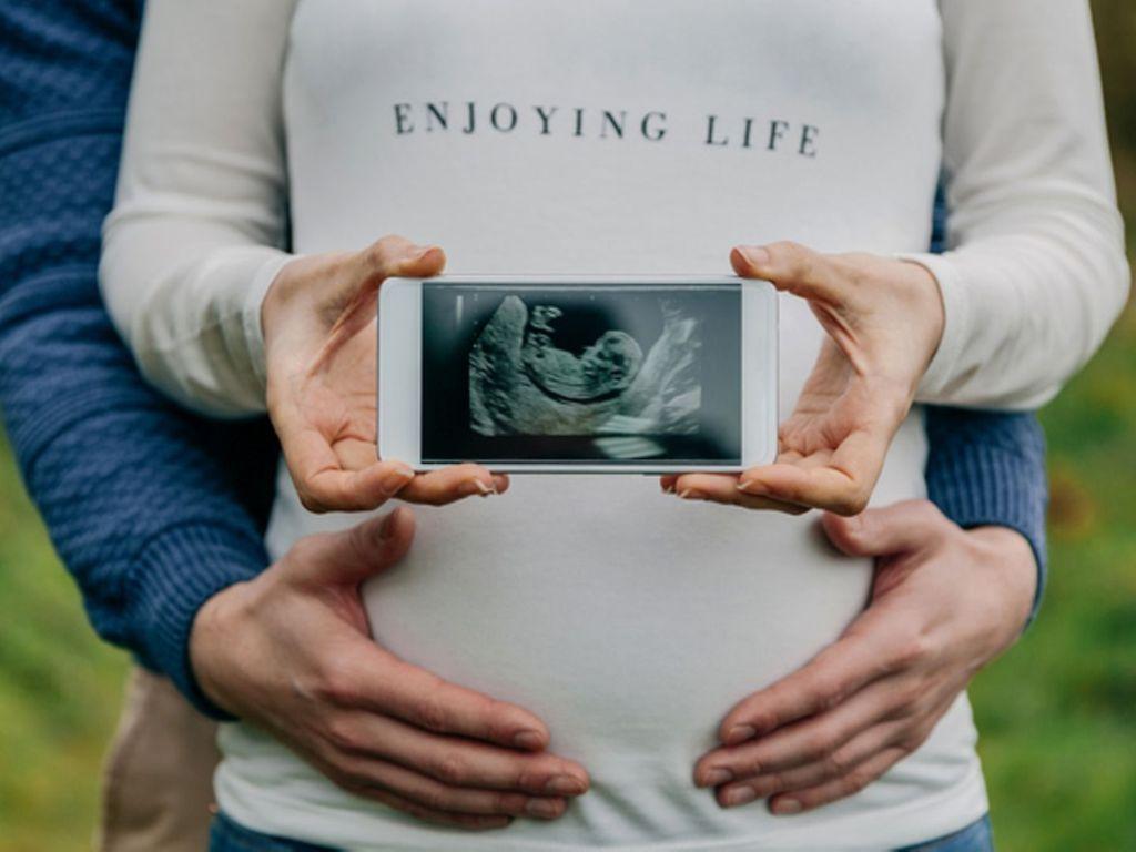 Schwangerschaft verkünden: Schwangere Frau, die Ultraschallbild in die Kamera hält und von ihrem Partner umfasst wird