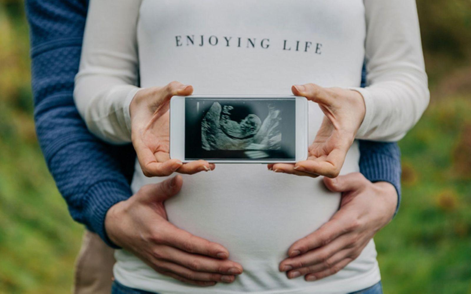 Schwangerschaft verkünden: Schwangere Frau, die Ultraschallbild in die Kamera hält und von ihrem Partner umfasst wird