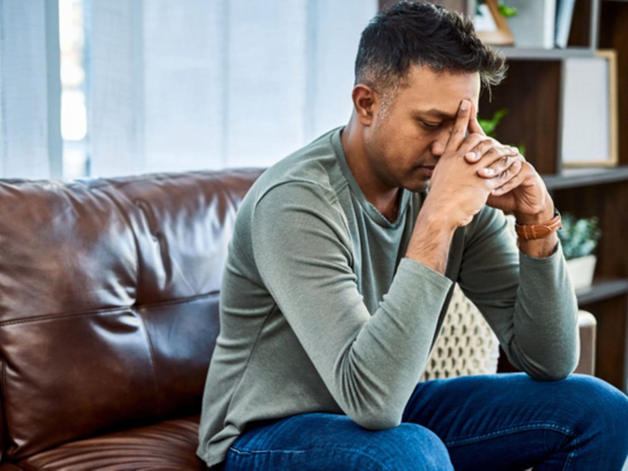 Depressionen: Mann sitzt verzweifelt auf der Couch mit seiner Stirn auf seine Hände gestützt