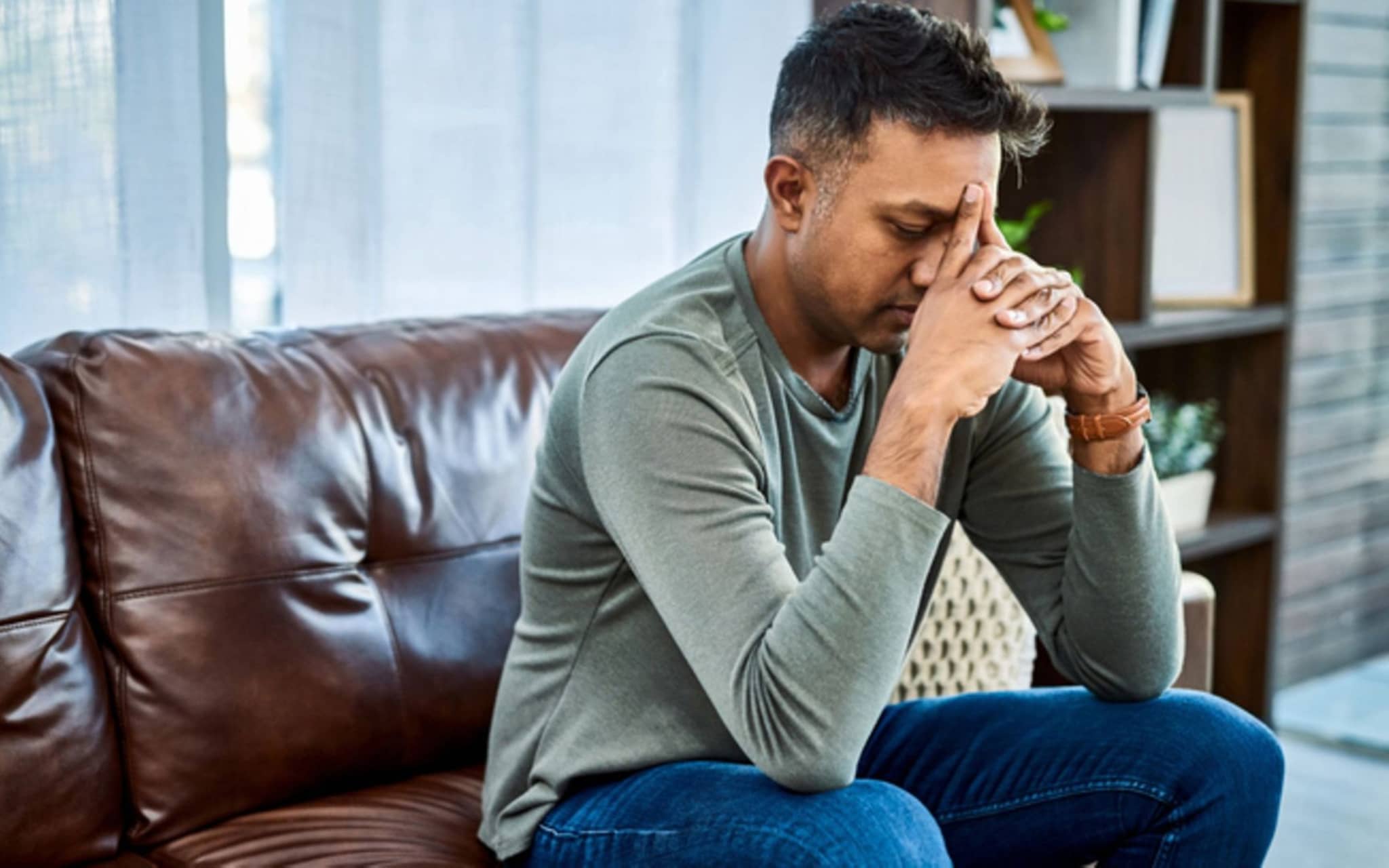 Depressionen: Mann sitzt verzweifelt auf der Couch mit seiner Stirn auf seine Hände gestützt