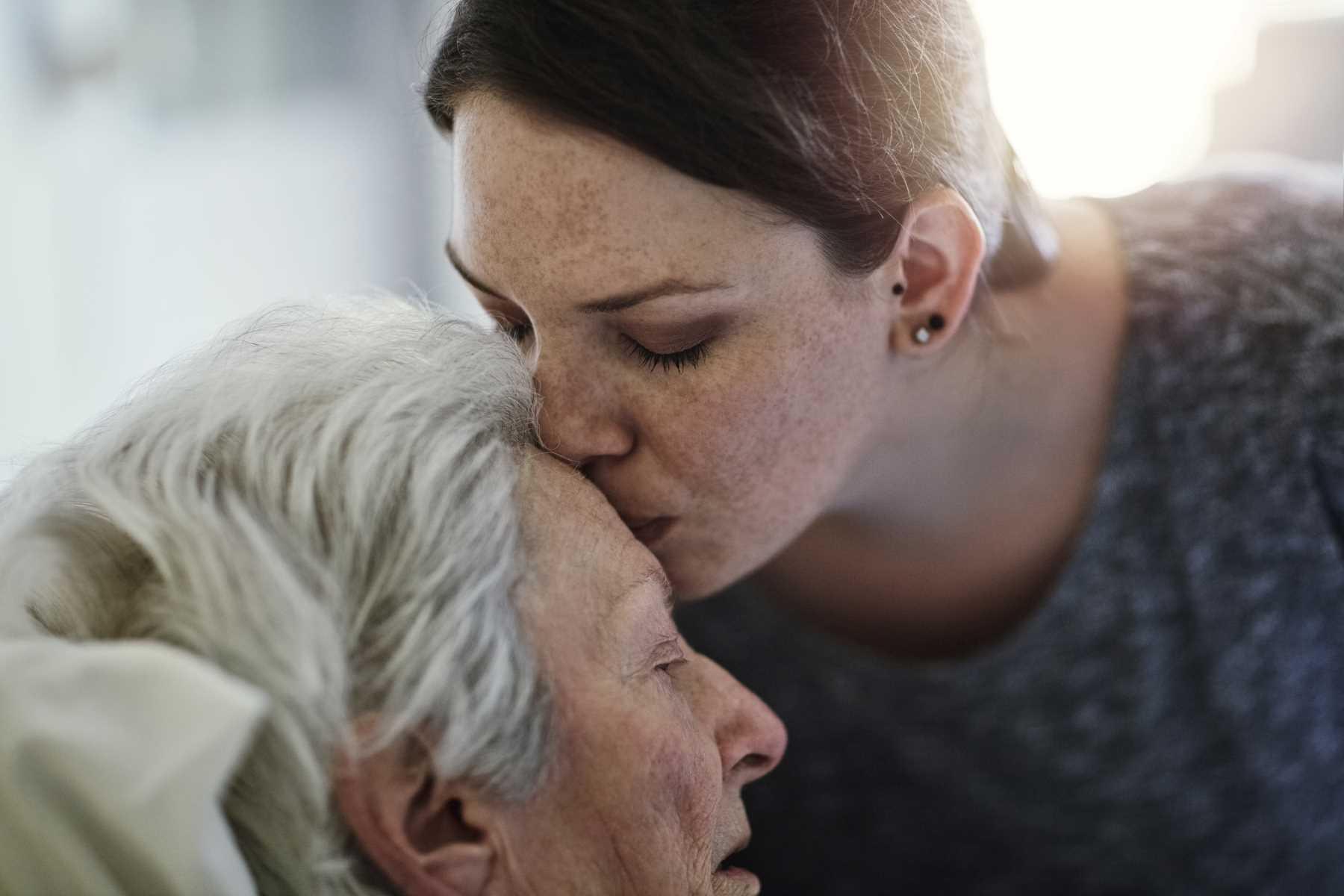 Zweitmeinung Intensiv: Jüngere Frau küsst ältere pflegebedürftige Frau auf die Stirn.