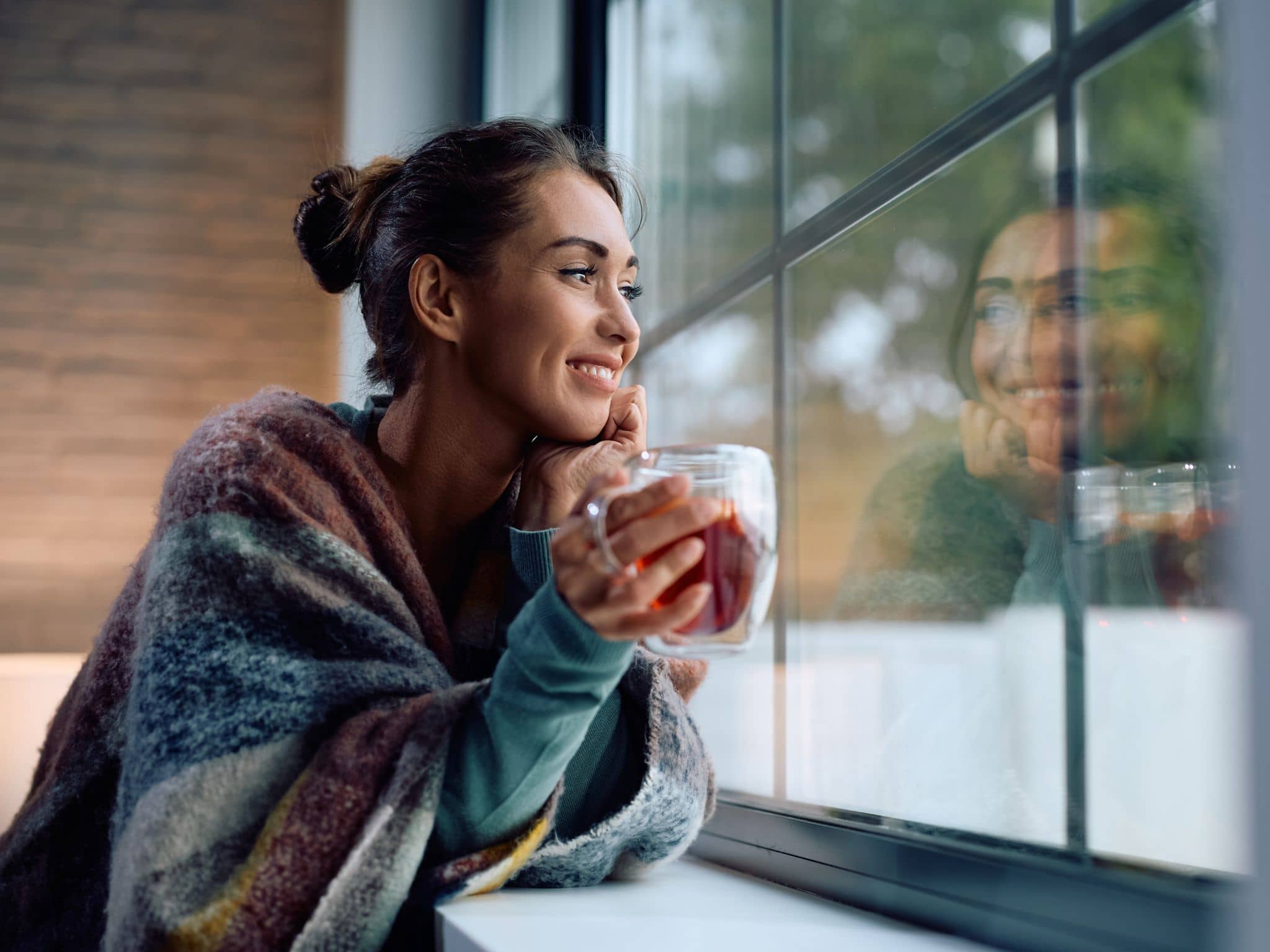 Heißgetränke: Frau mit Tasse in der Hand schaut lächelnd aus dem Fenster.