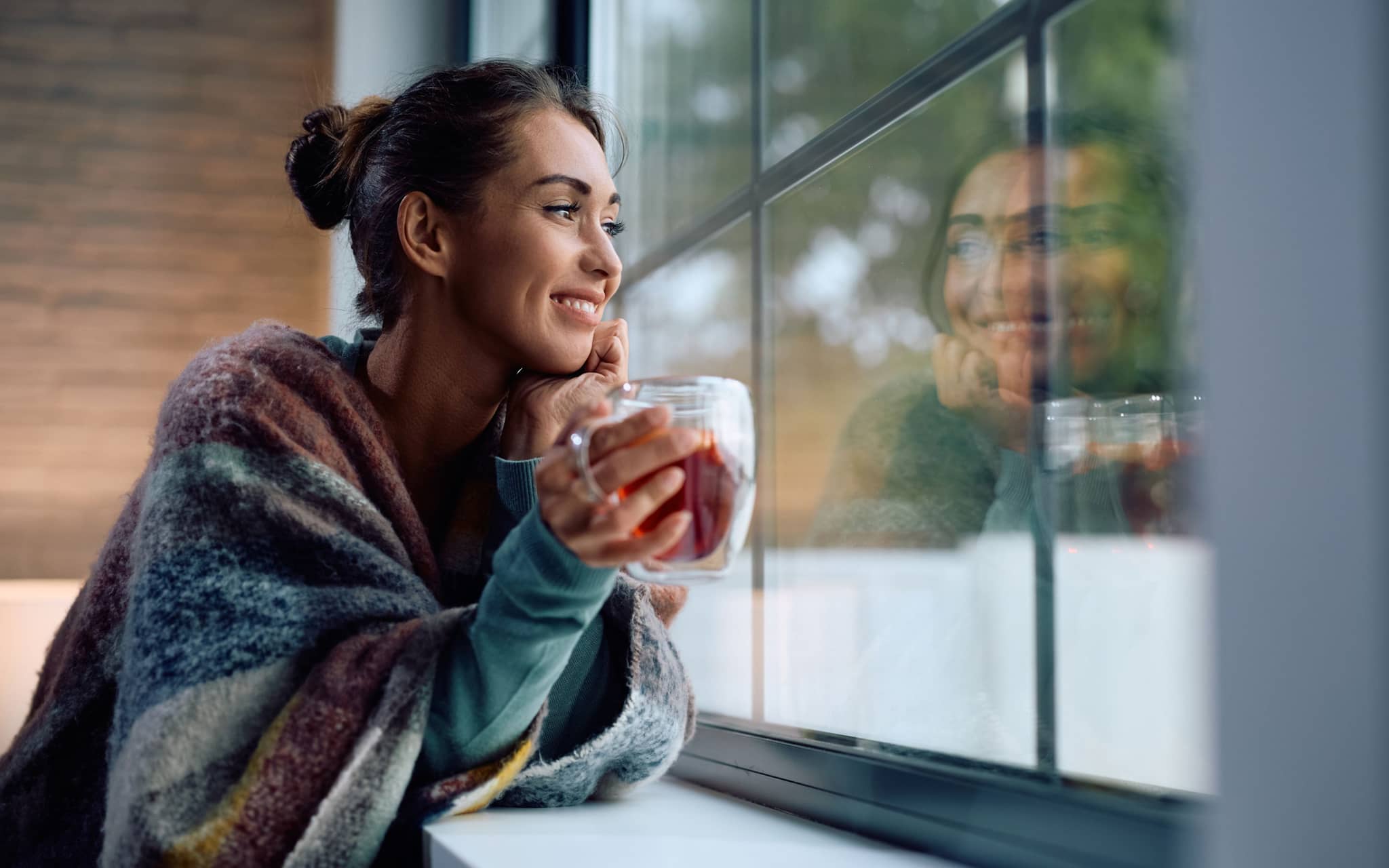 Heißgetränke: Frau mit Tasse in der Hand schaut lächelnd aus dem Fenster.
