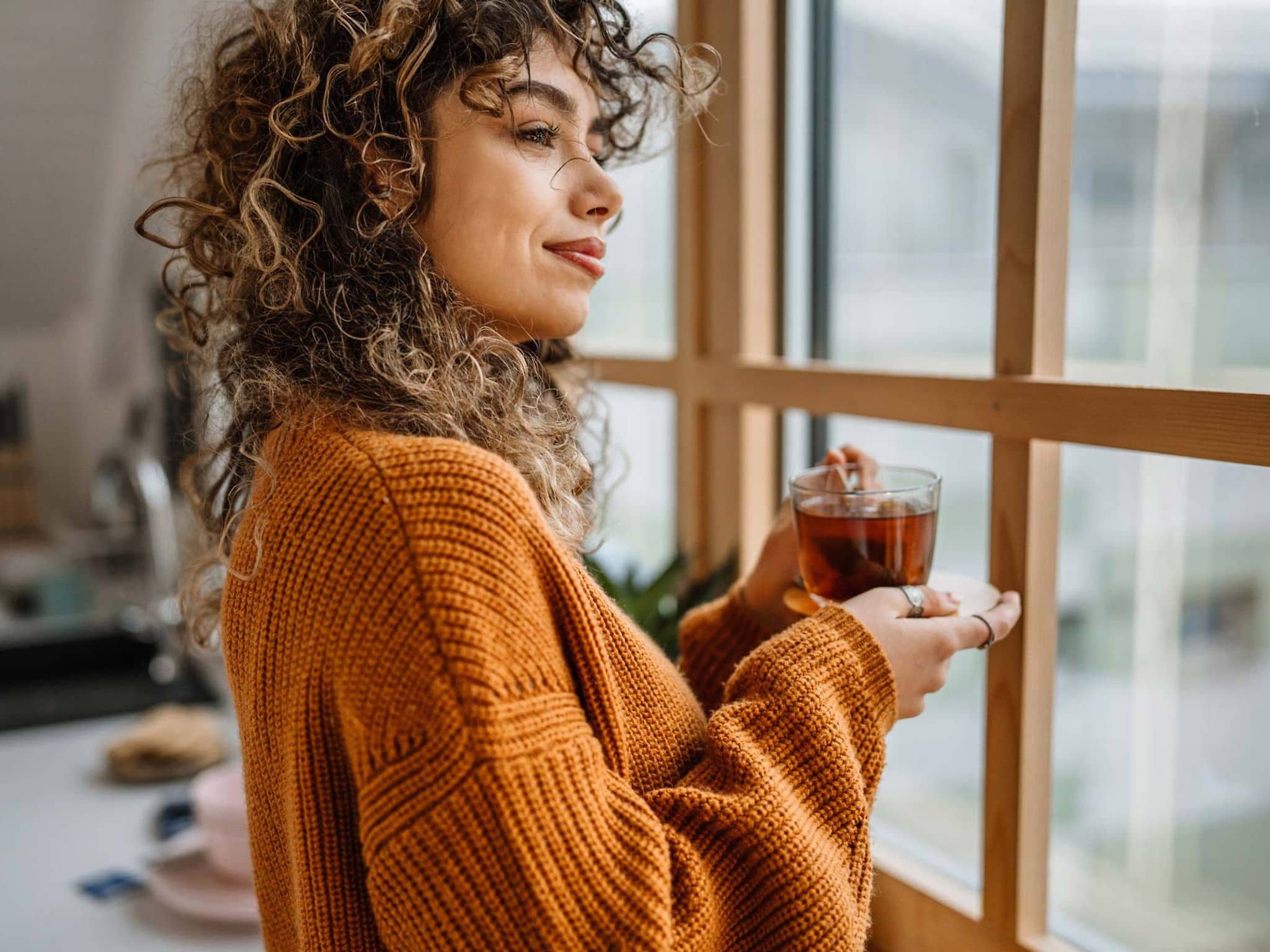 Tee ist gesund: Junge Frau steht mit einer Tasse Tee in der Küche und schaut aus dem Fenster.