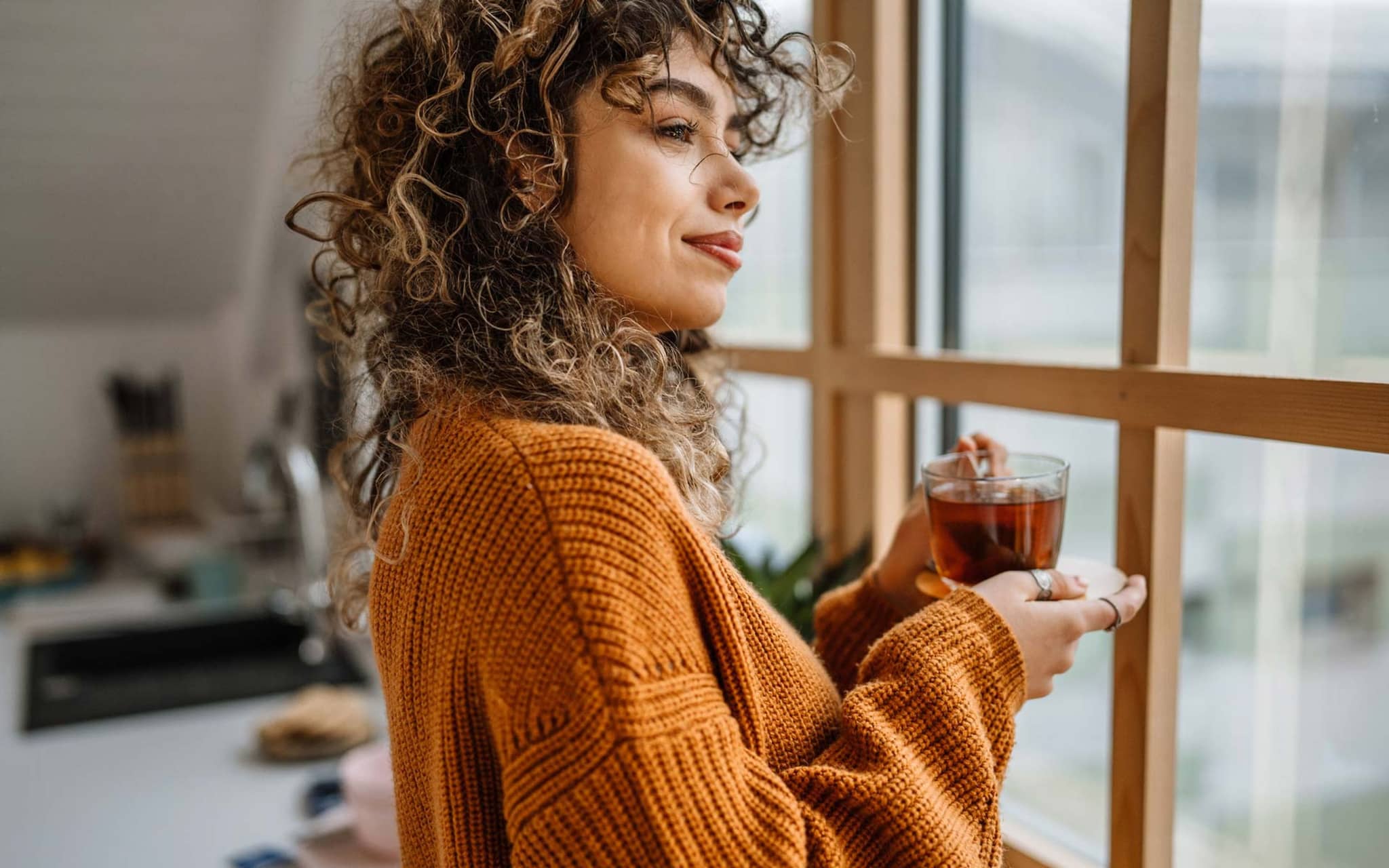 Tee ist gesund: Junge Frau steht mit einer Tasse Tee in der Küche und schaut aus dem Fenster.