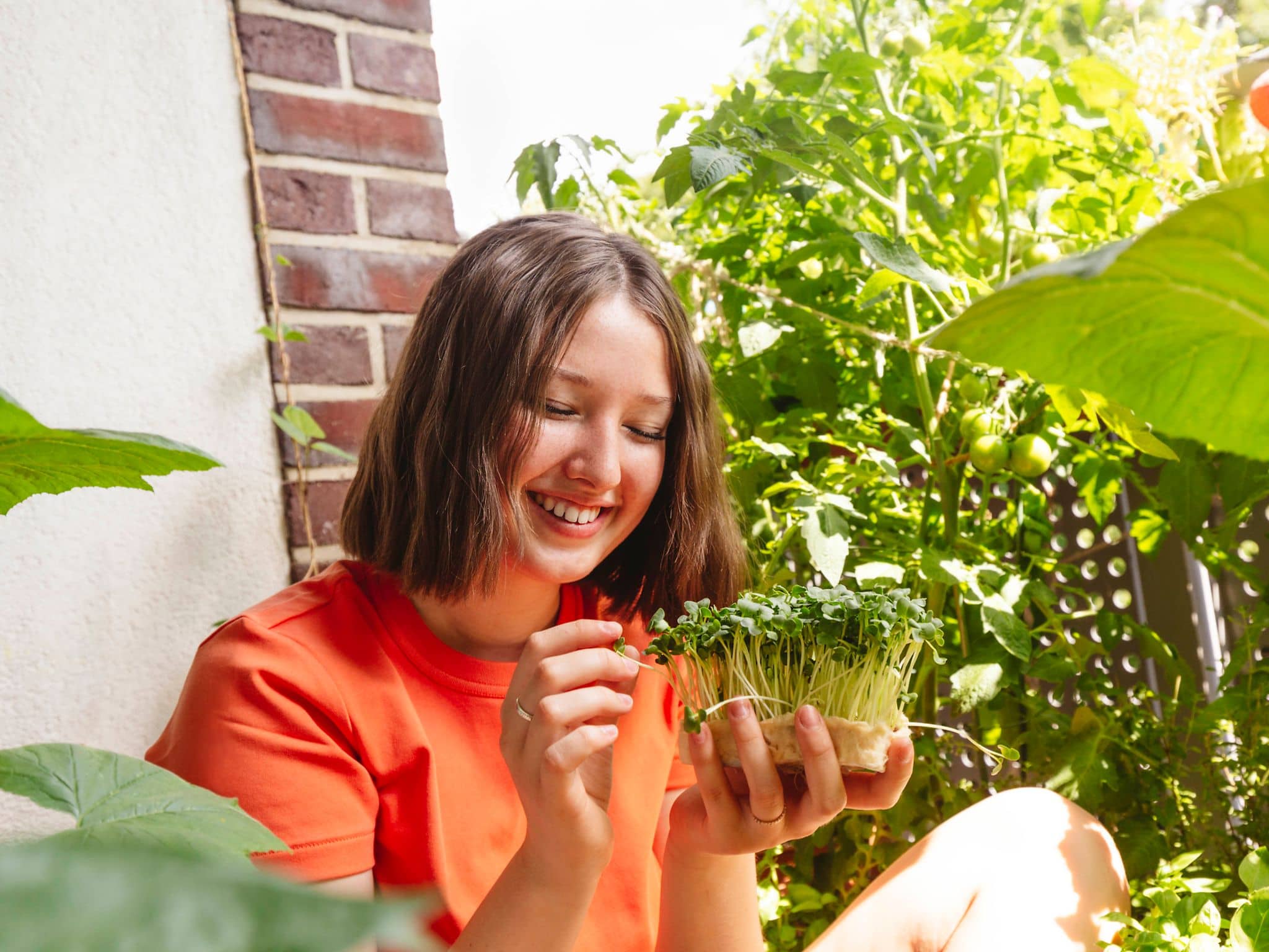 Junge Frau sitzt auf dem Balkon und betrachtet lachend die gesunden Microgreens in ihrer Hand.