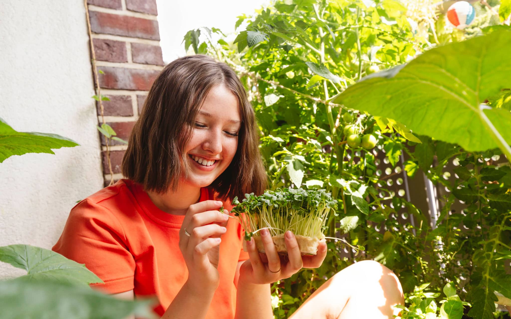 Junge Frau sitzt auf dem Balkon und betrachtet lachend die gesunden Microgreens in ihrer Hand.