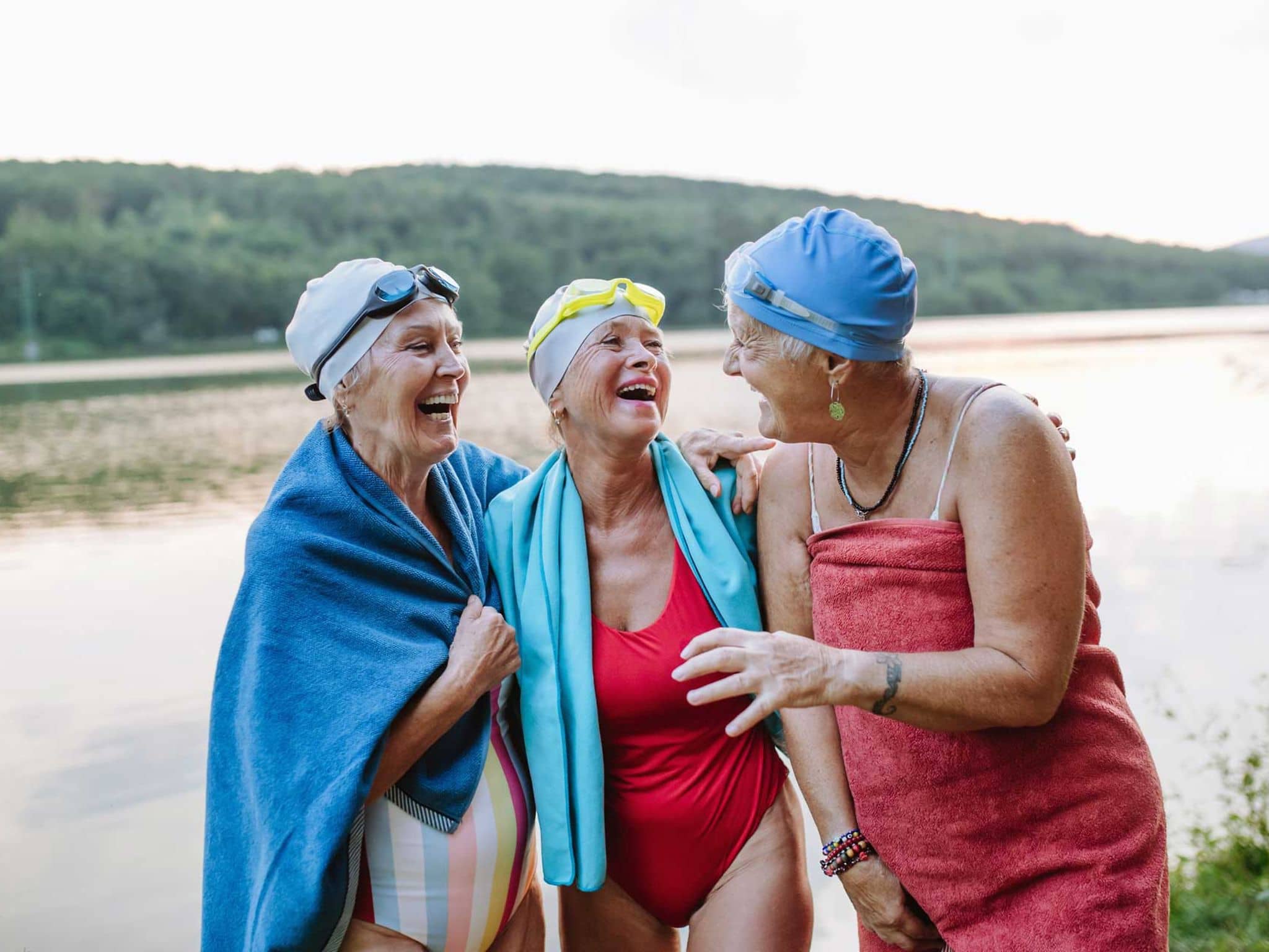 Drei ältere Frauen stehen lachend in Badekleidung vor einem See