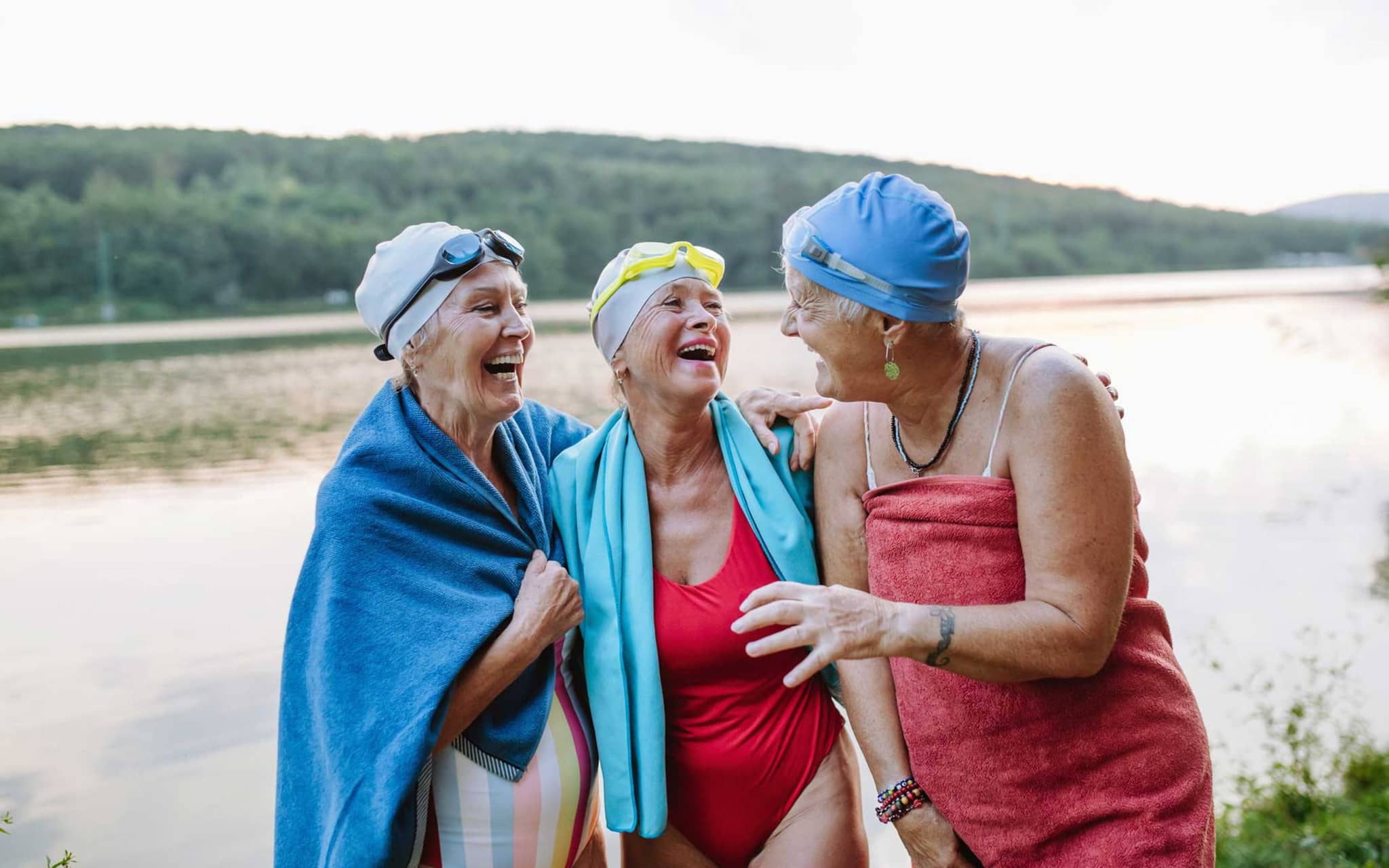 Drei ältere Frauen stehen lachend in Badekleidung vor einem See