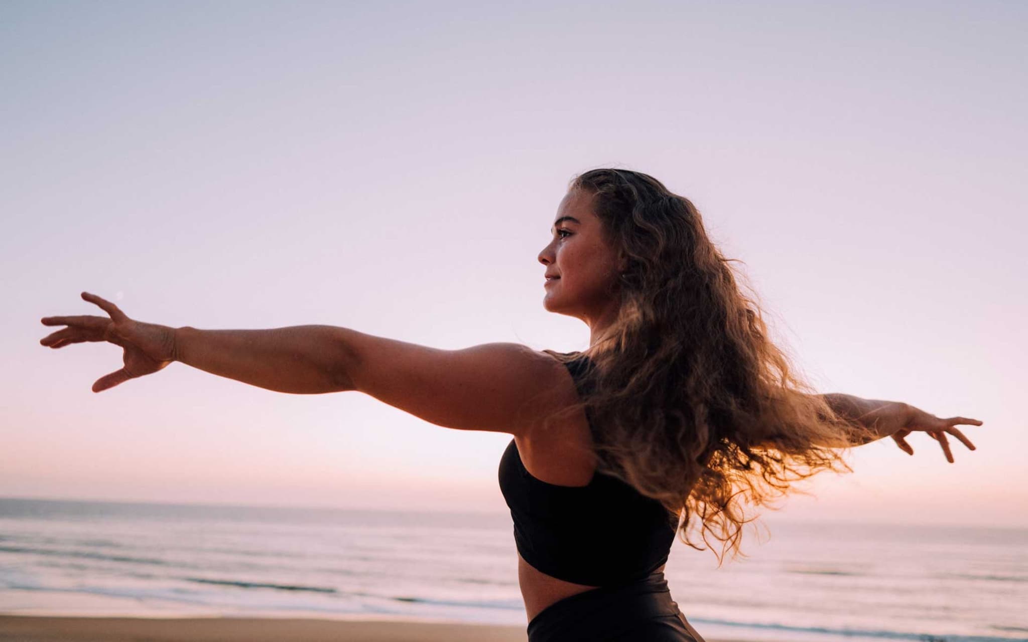 Junge Frau blickt fröhlich mit ausgebreiteten Armen über den Strand