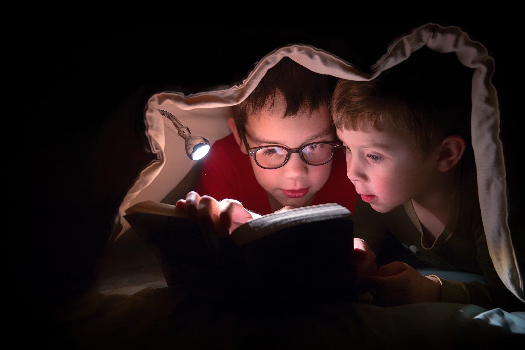 Zwei Kinder lesen nachts unter einer Decke in einem Buch
