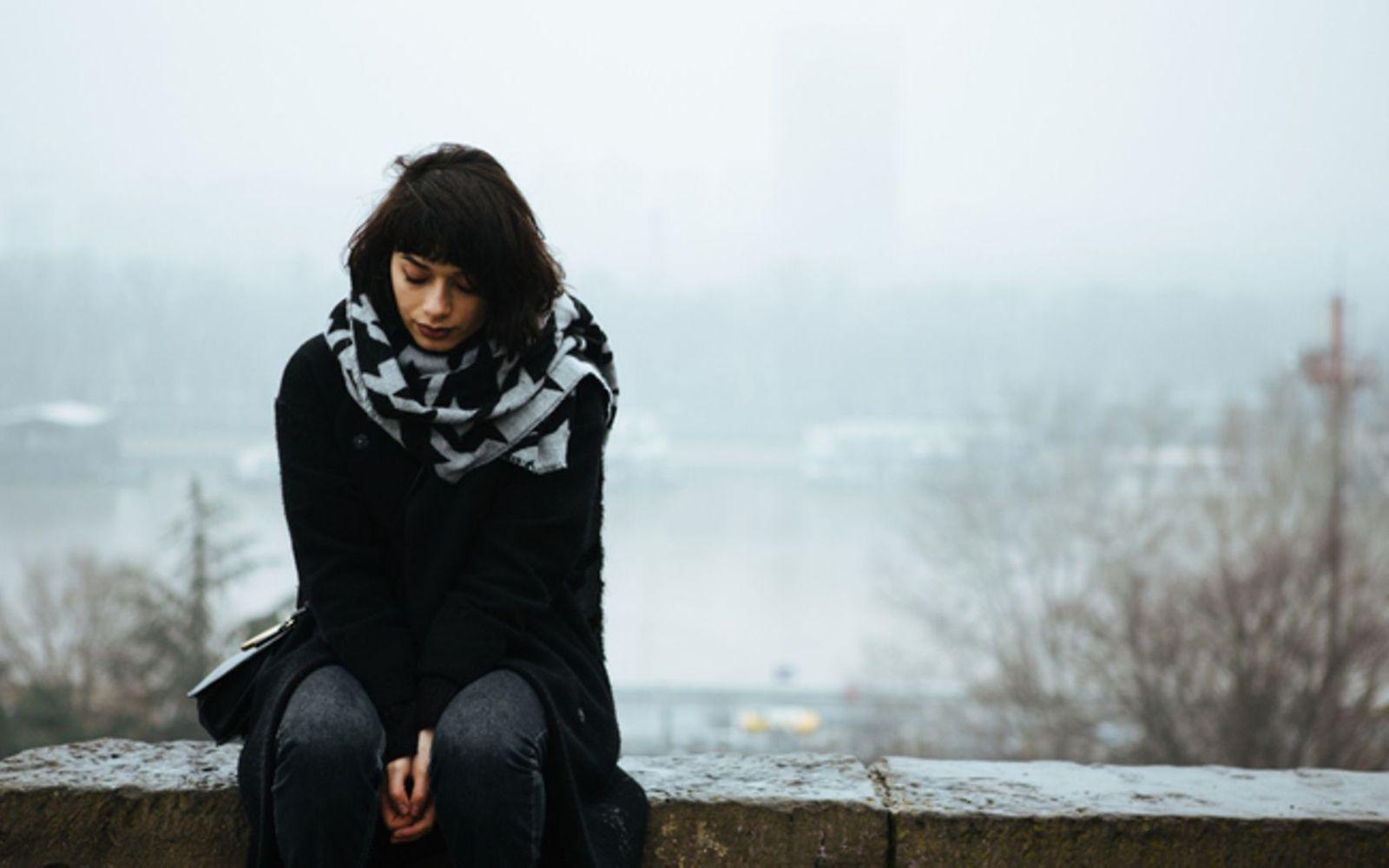 Winterdepression: Frau sitzt in einer Winterlandschaft traurig auf einer Mauer.
