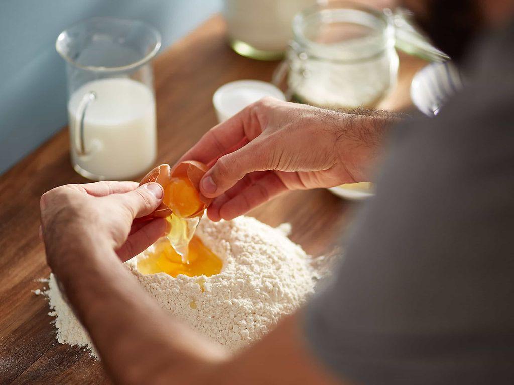Lebensmittelallergie: Zwei Hände geben ein aufgeschlagenes Ei in eine Mehlmulde.
