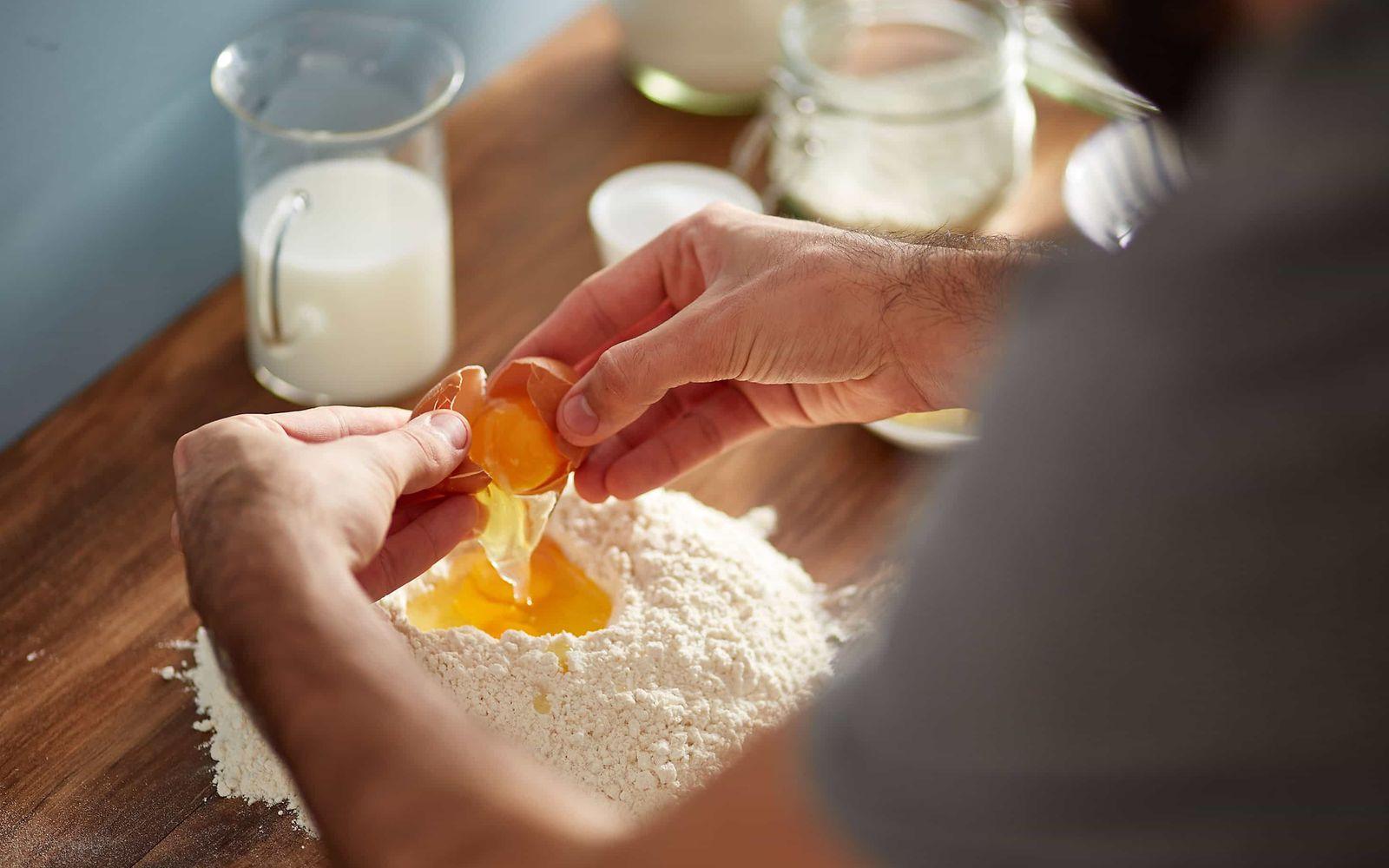 Lebensmittelallergie: Zwei Hände geben ein aufgeschlagenes Ei in eine Mehlmulde.