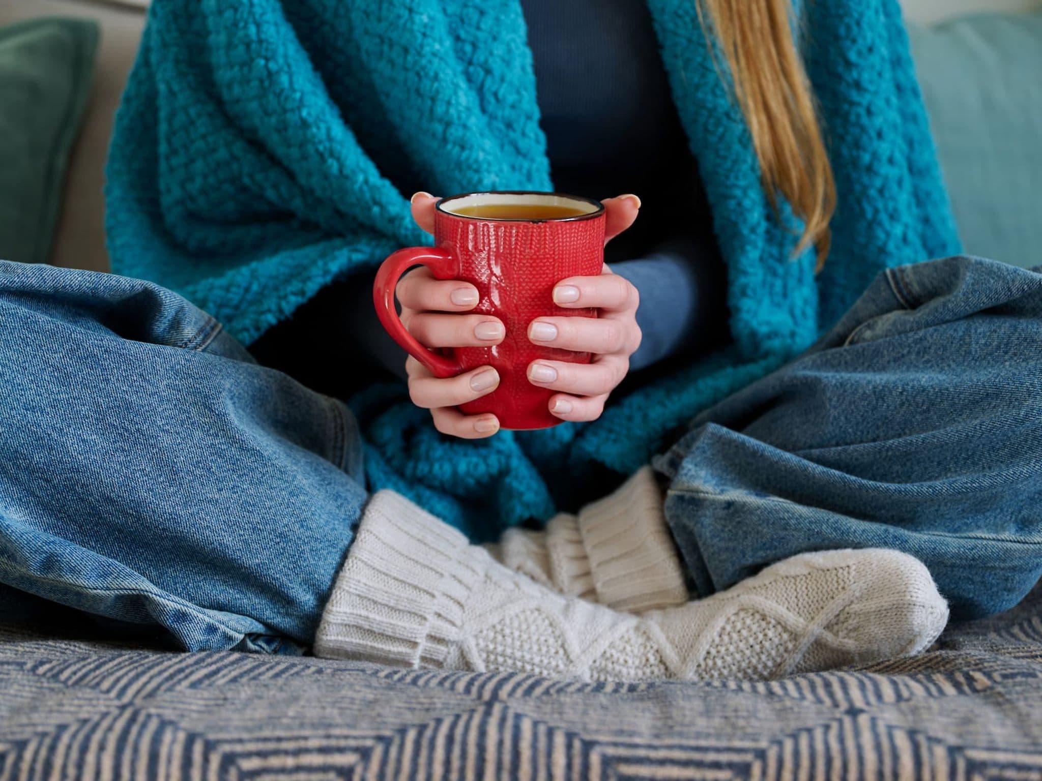 Frau sitzt im Schneidersitz und wärmt ihre Hände mit einer Tasse und ihre Füße mit Wollsocken