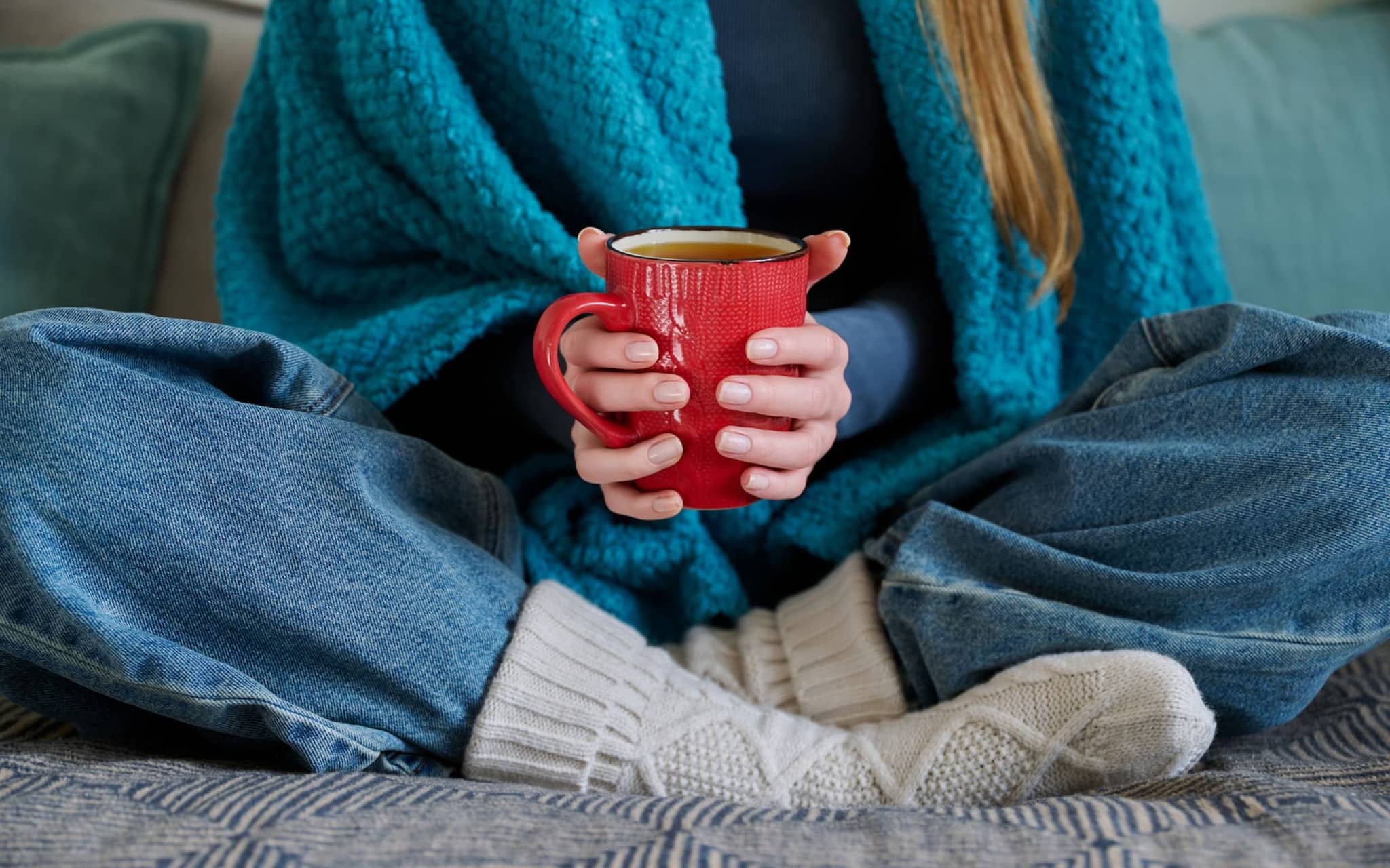 Frau sitzt im Schneidersitz und wärmt ihre Hände mit einer Tasse und ihre Füße mit Wollsocken