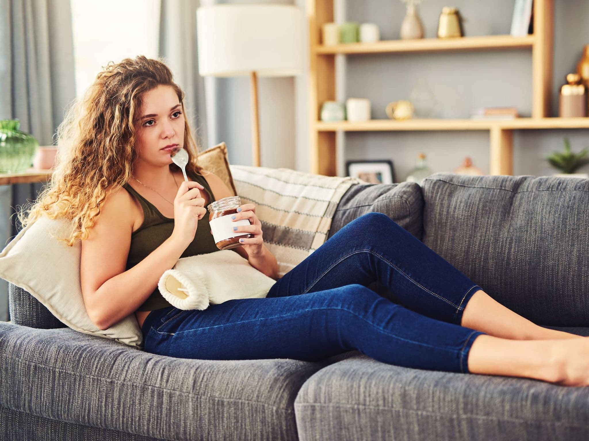Betrübte, junge Frau sitzt auf dem Sofa mit Wärmflasche und isst mit dem Löffel Schokoaufstrich aus einem Glas