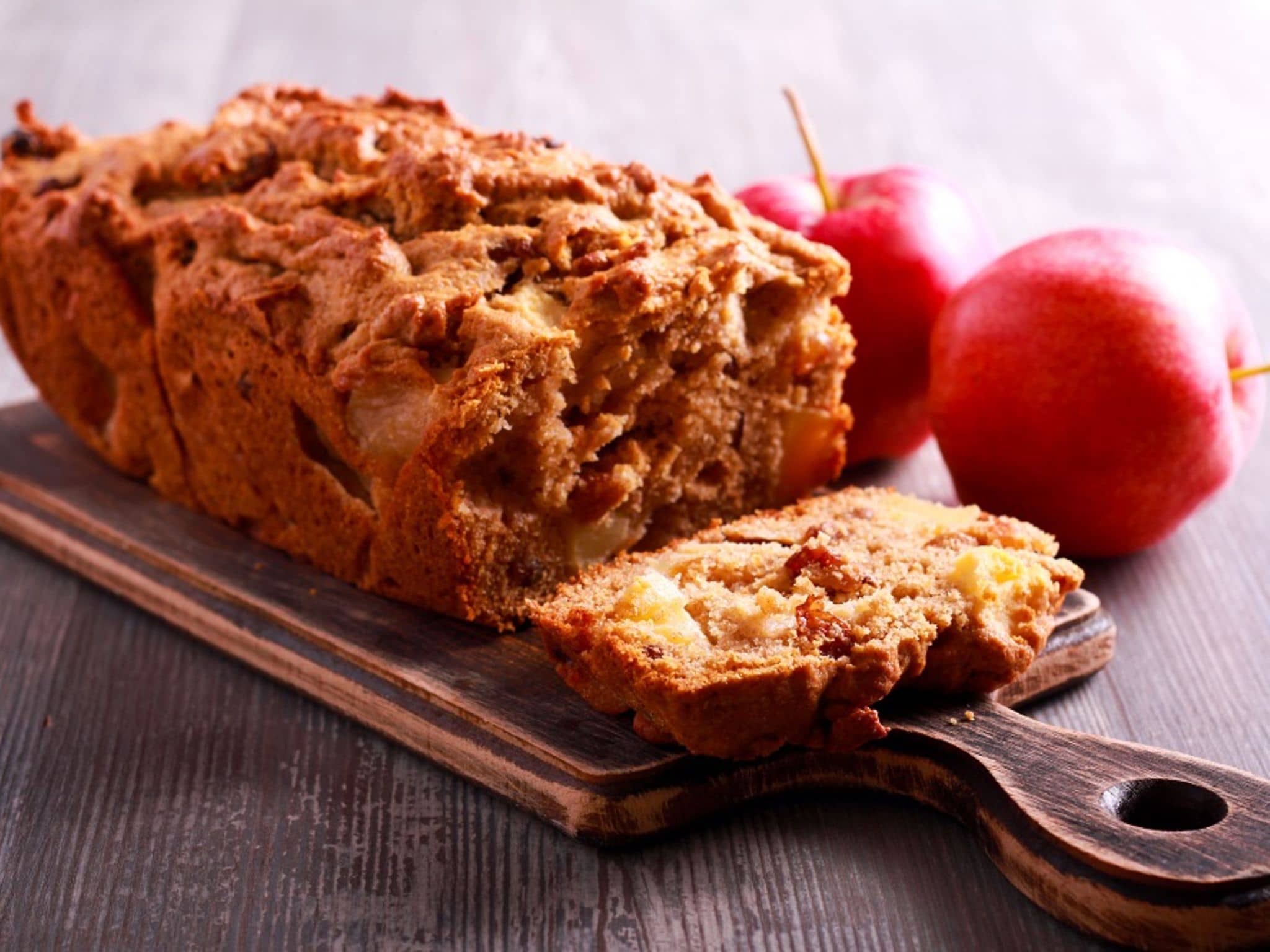 Frisch angeschnittenes Apfelbrot auf einem Brett drapiert. Daneben liegen zwei Äpfel.
