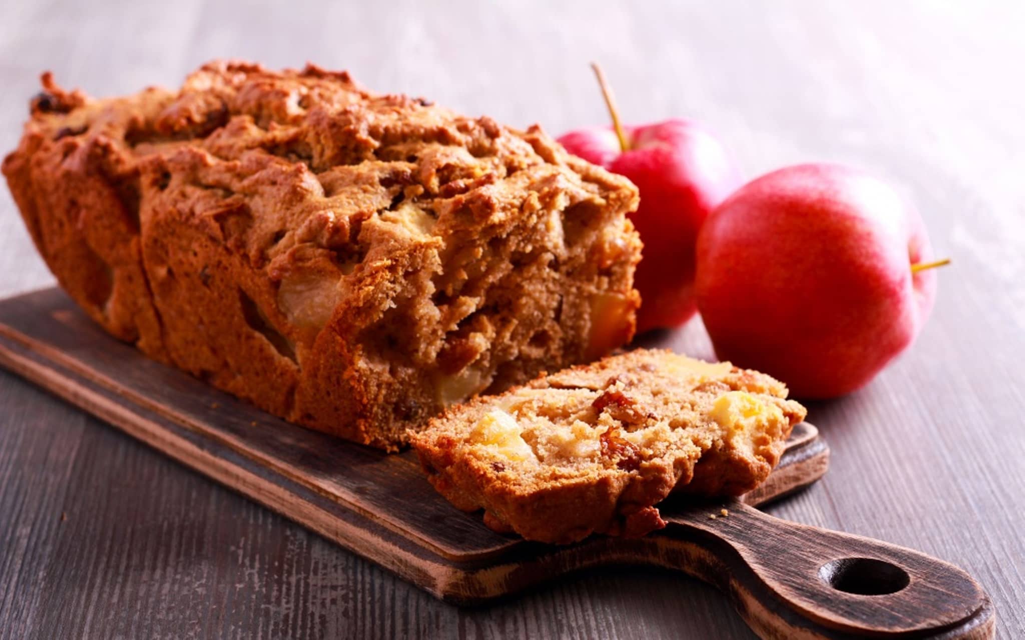 Frisch angeschnittenes Apfelbrot auf einem Brett drapiert. Daneben liegen zwei Äpfel.