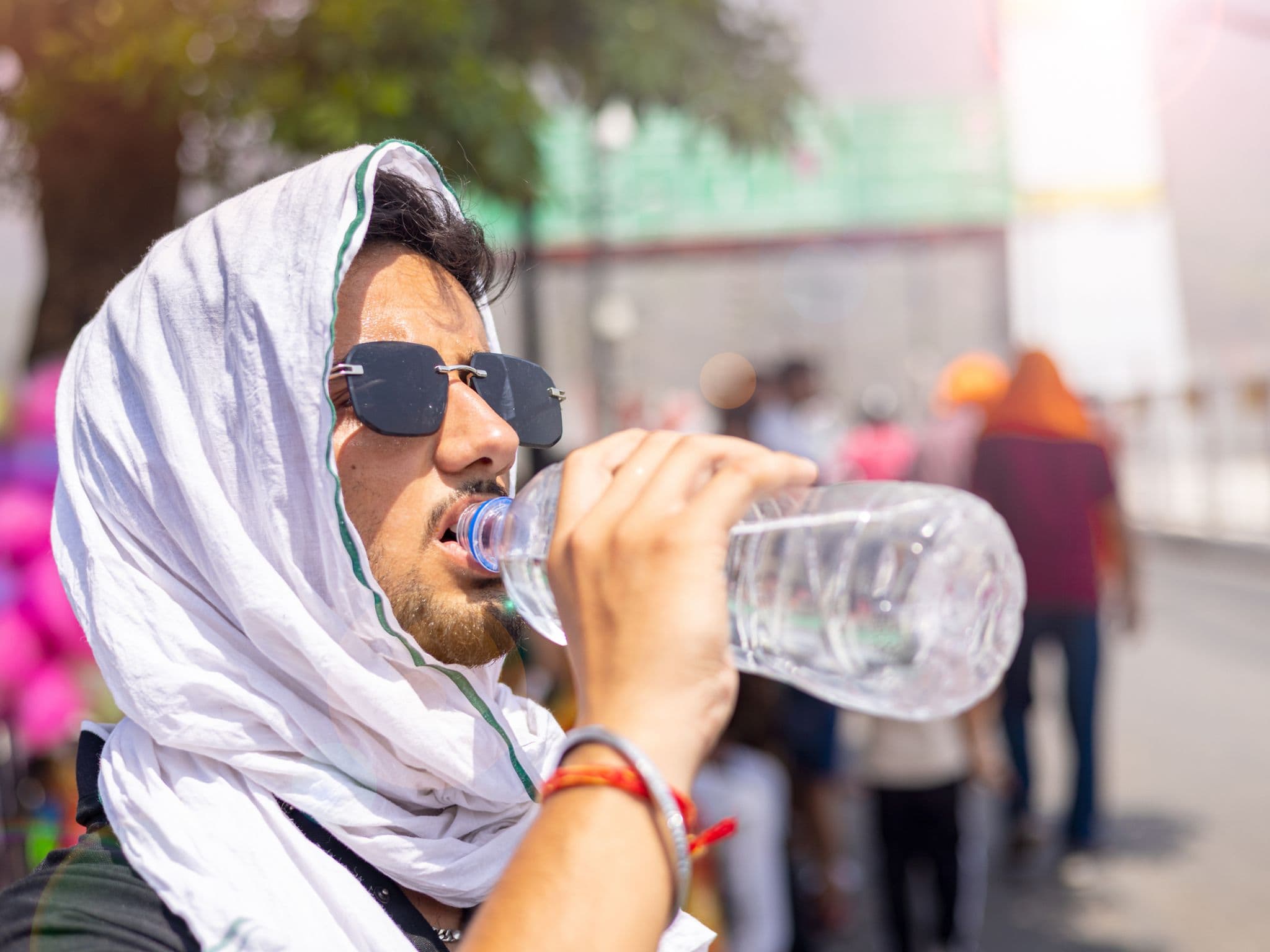 Tipps bei Hitze: Junger Mann schützt sich vor Sonne und Hitze mit einem weißen Kopftuch und trinkt Wasser aus einer Flasche.