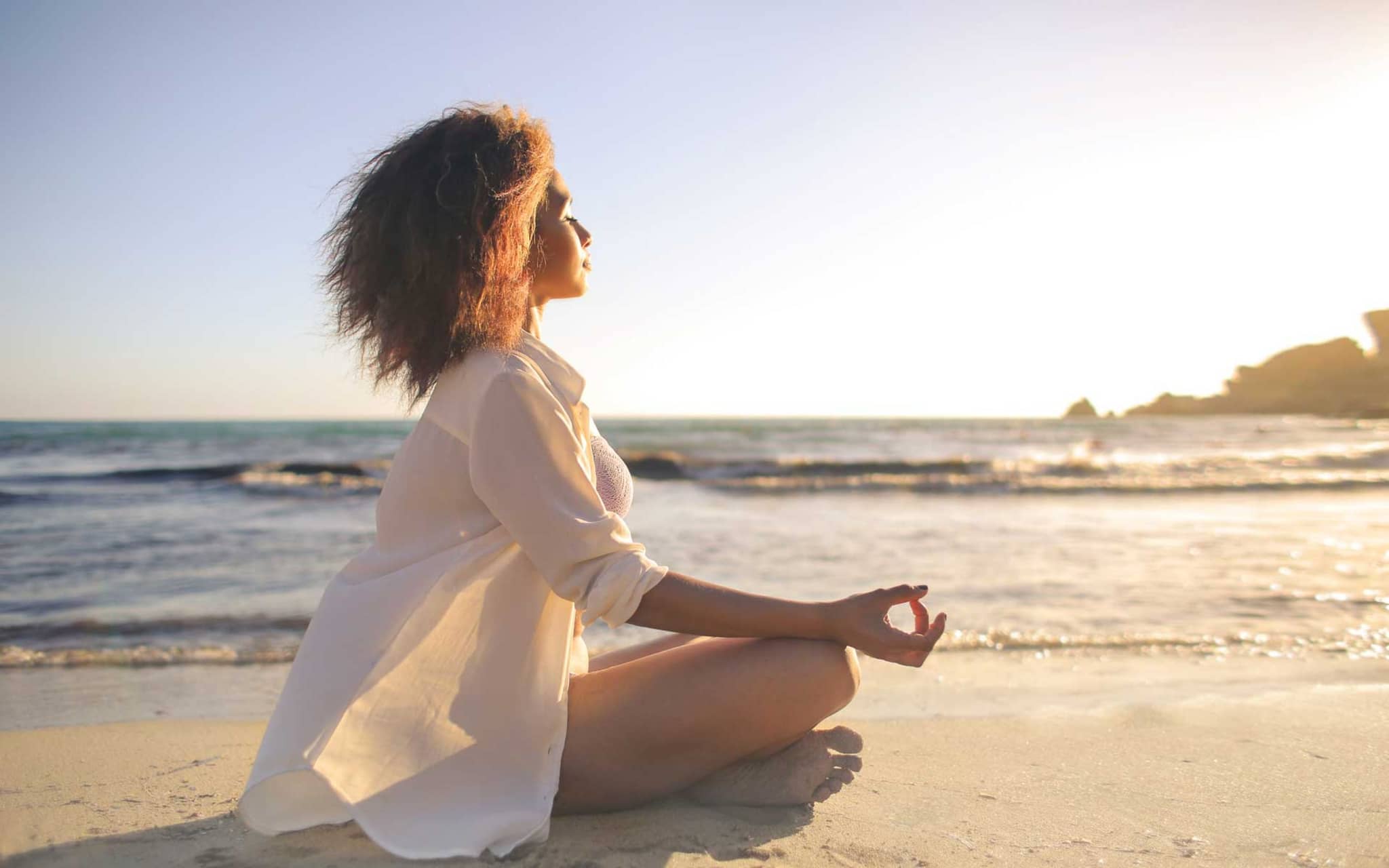 Frau sitzt meditierend am Strand und genießt die warme Meeresbrise