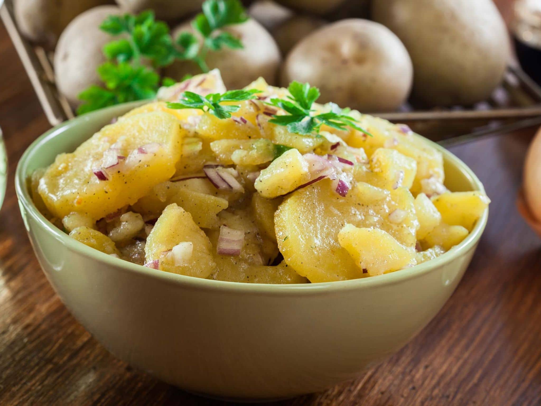 Rezeptfoto: Schüssel mit Kartoffelsalat. Daneben liegen rohe Kartoffeln.