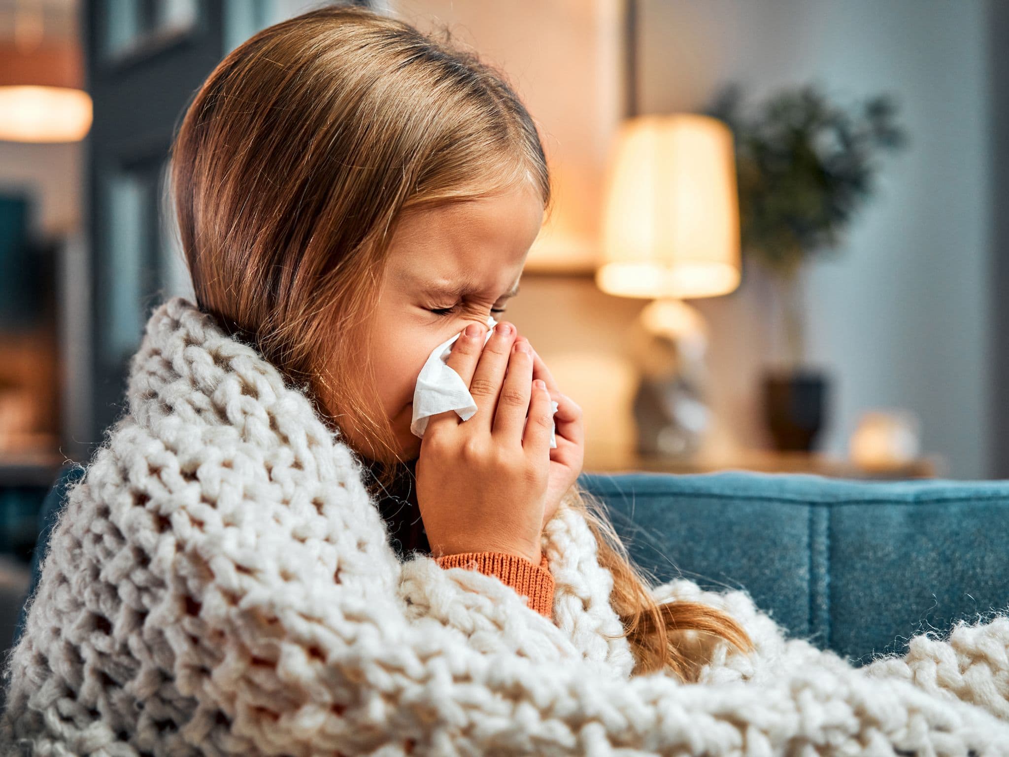 Erkältung bei Kindern: Kleines Mädchen auf dem Sofa putzt sich mit einem Taschentuch die Nase.