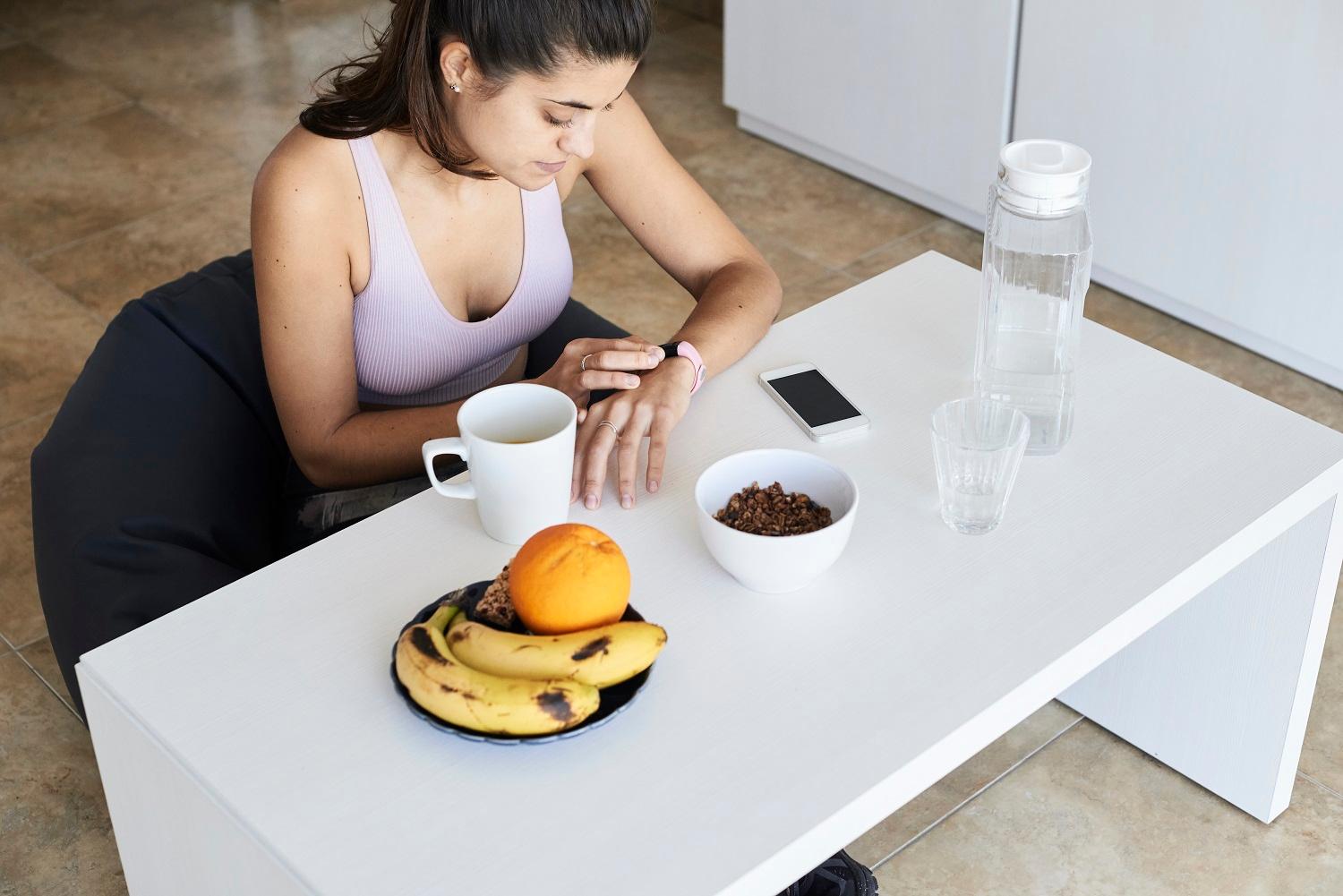 Mehr Kick durch Kaffee: Junge Frau sitzt am Esstisch und tippt auf ihre Smartwatch