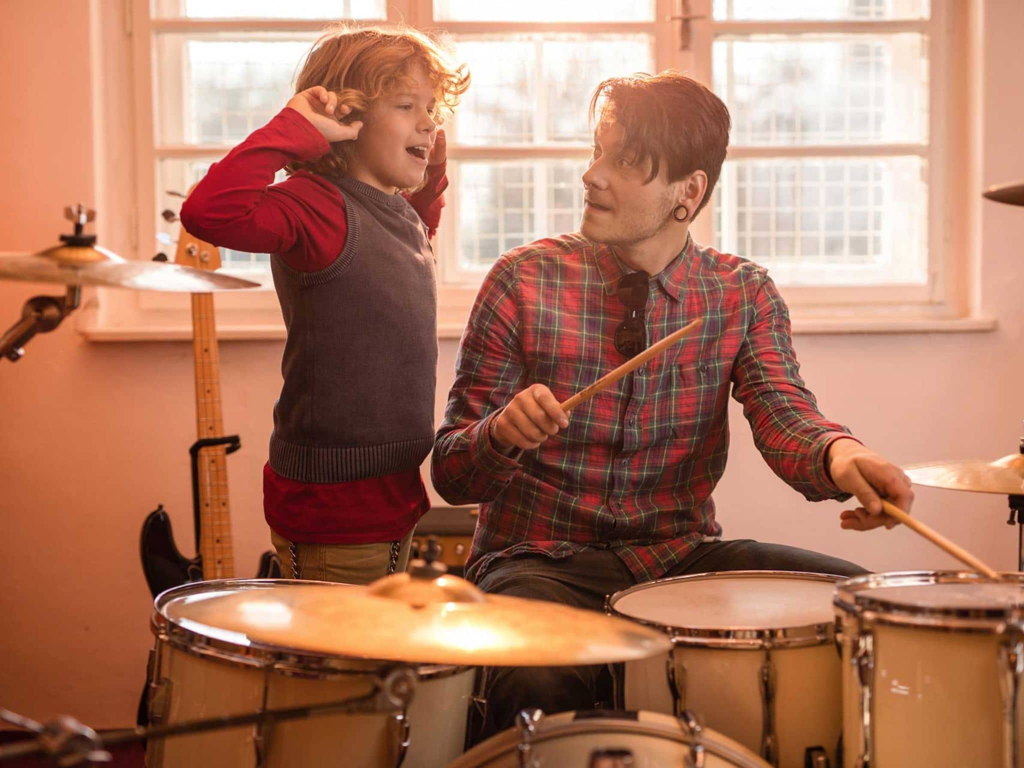 Junge hält sich die Ohren zu, während Vater Schlagzeug spielt