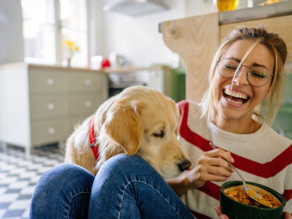 Vorbeugung von Parodontitis: Junge Frau sitzt lachend mit ihrem Hund auf dem Boden mit einer Schale Essen in der Hand.