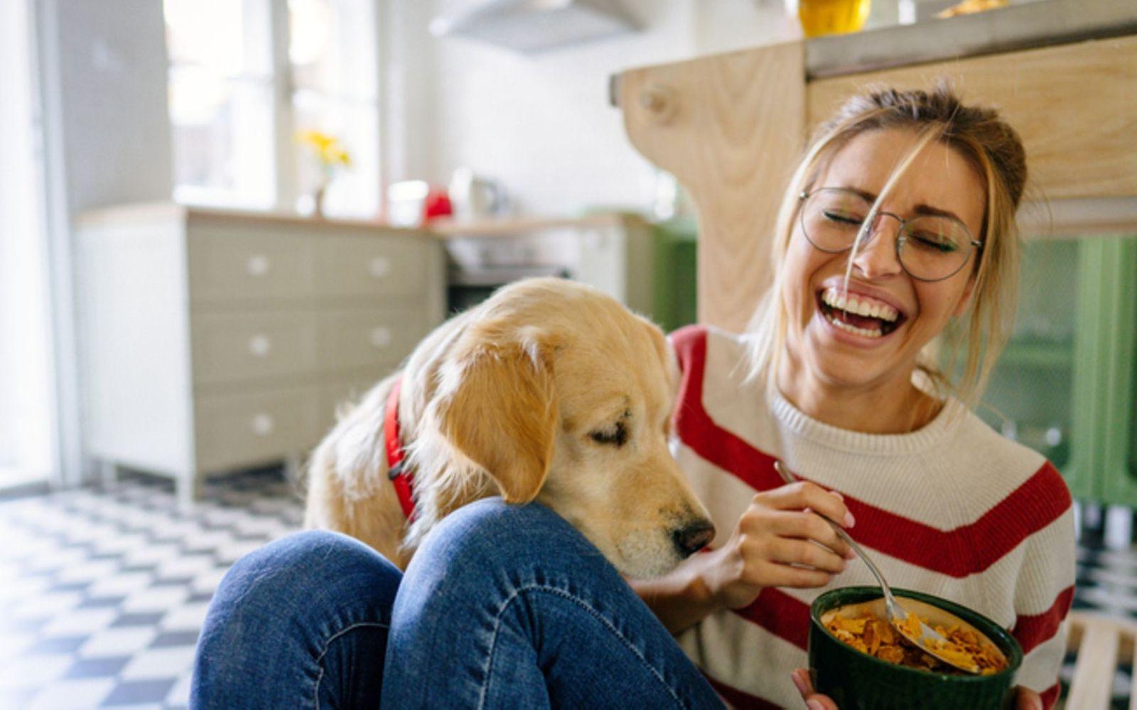 Vorbeugung von Parodontitis: Junge Frau sitzt lachend mit ihrem Hund auf dem Boden mit einer Schale Essen in der Hand.