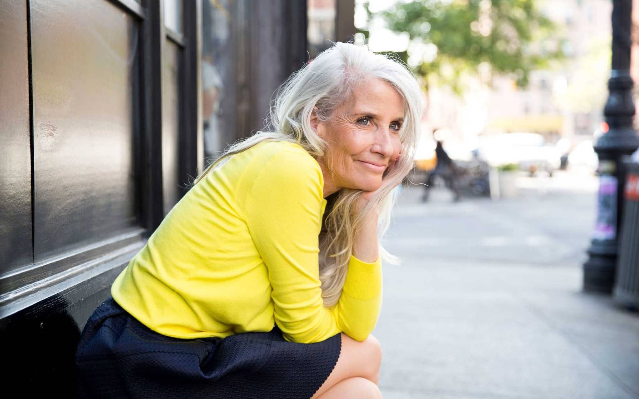 Wechseljahresbeschwerden: Ältere, draußen sitzende Frau stützt lächelnd den Kopf auf die Hand.