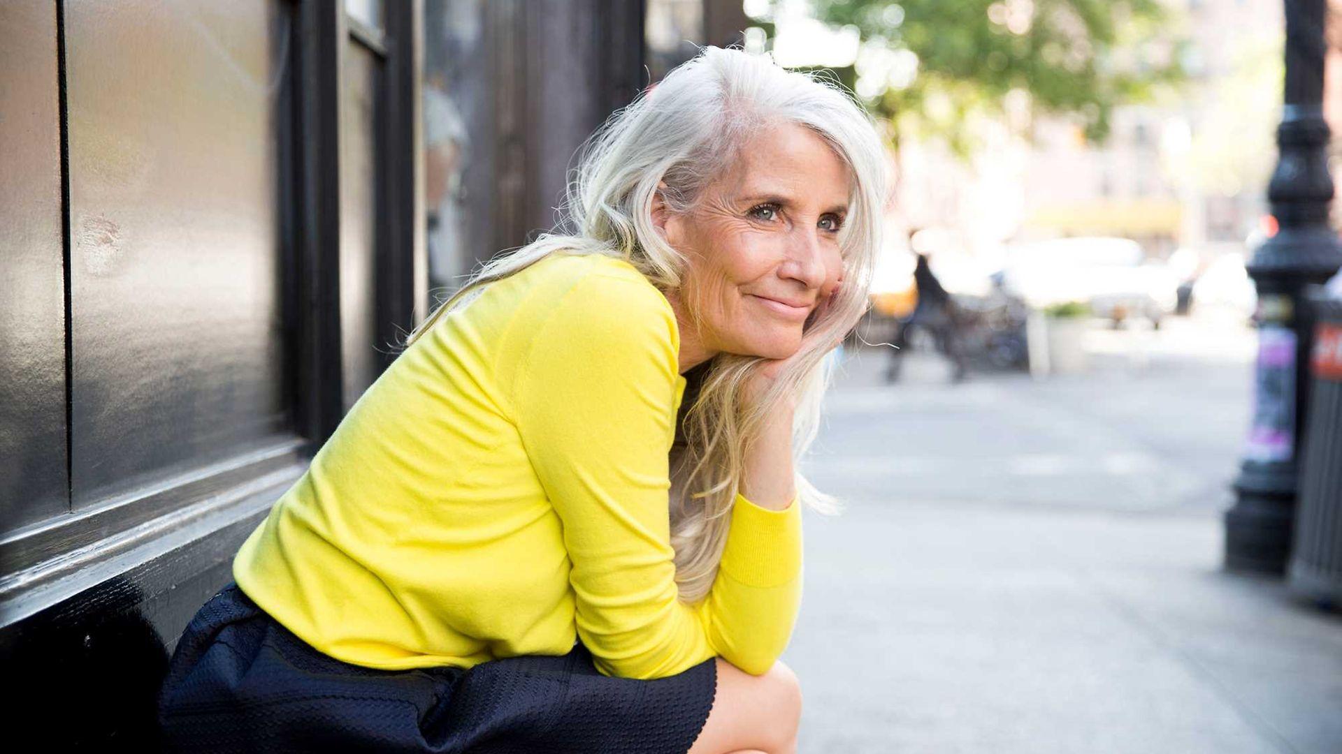 Wechseljahresbeschwerden: Ältere, draußen sitzende Frau stützt lächelnd den Kopf auf die Hand. 
