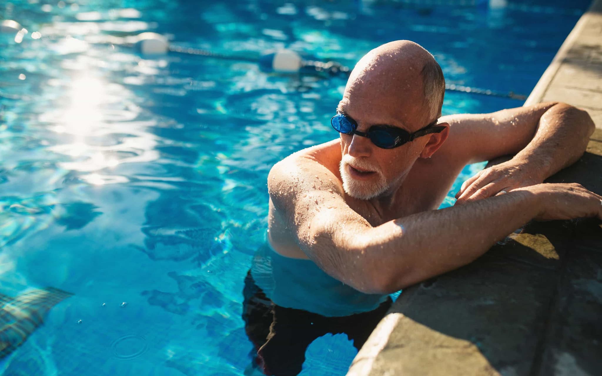 Herzinfarkt: Älterer Mann mit Schwimmbrille hält sich am Beckenrand eines Schwimmbeckens fest.