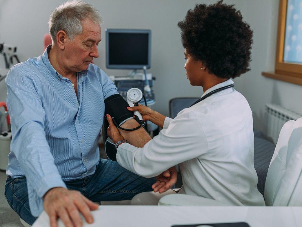 Niedriger Blutdruck: Ärztin misst bei älterem, auf einer Liege sitzenden Mann Blutdruck.