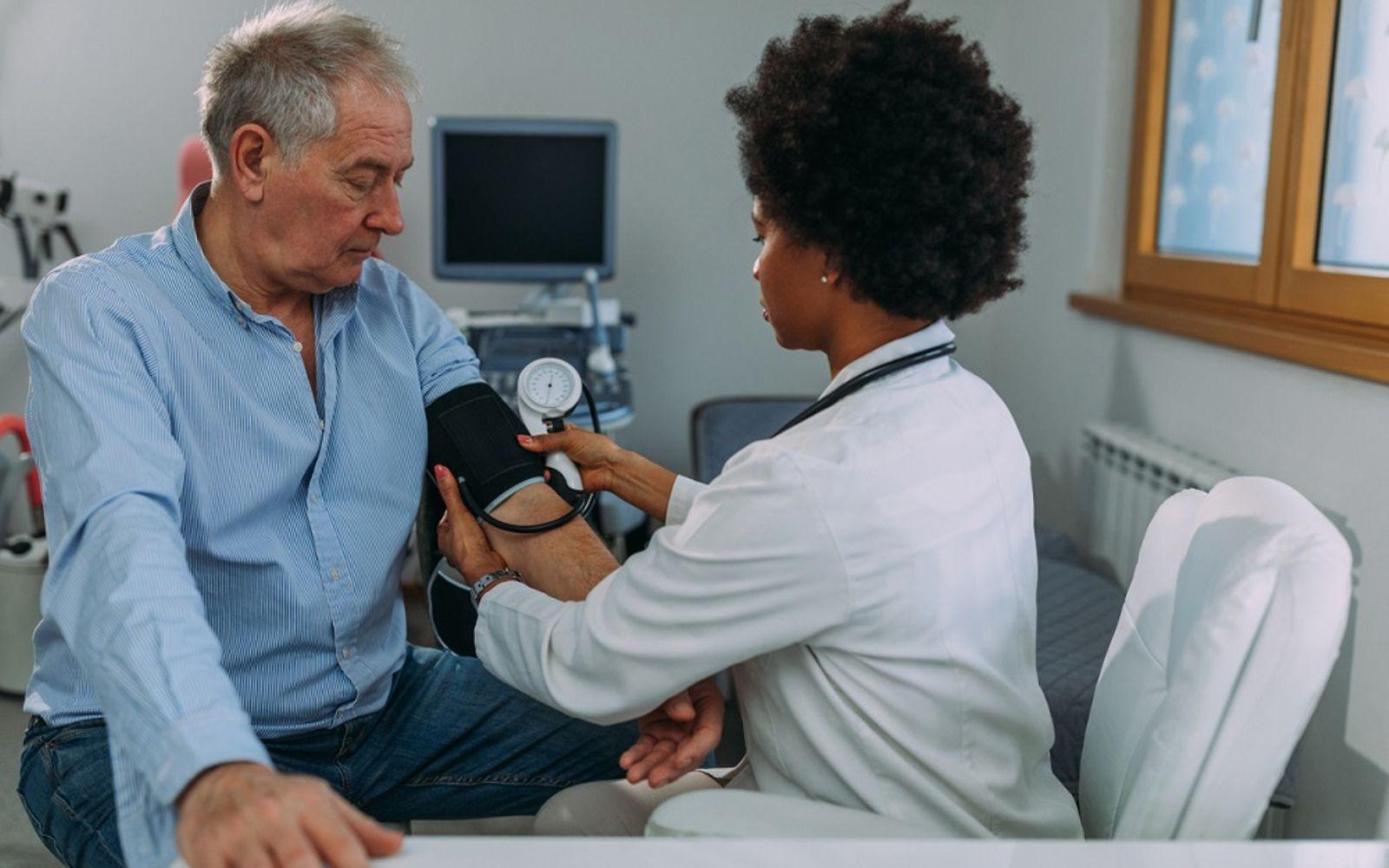 Niedriger Blutdruck: Ärztin misst bei älterem, auf einer Liege sitzenden Mann Blutdruck.