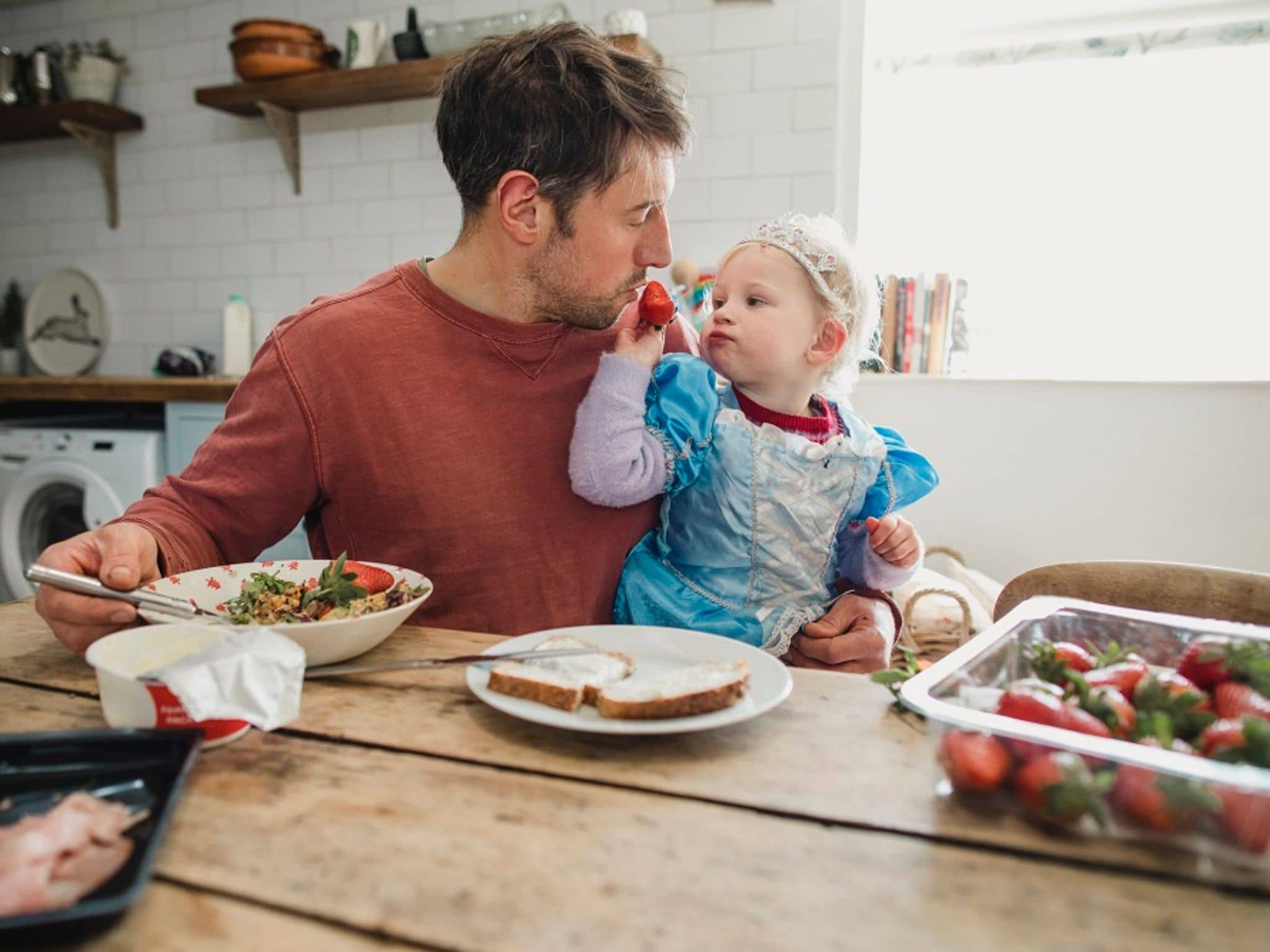 Ernährung bei Neurodermitis: Vater und Kleinkind am Küchentisch - Kind füttert Vater mit einer Erdbeere.