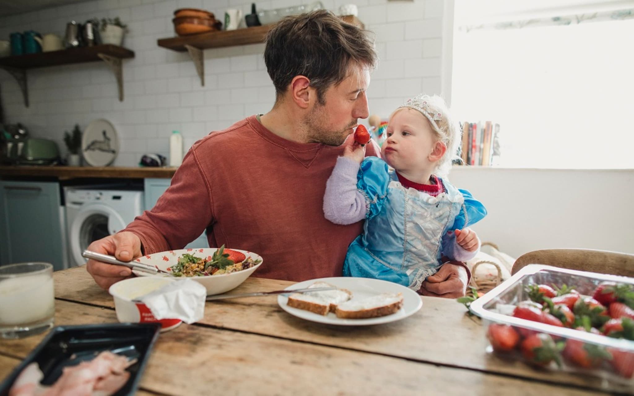 Ernährung bei Neurodermitis: Vater und Kleinkind am Küchentisch - Kind füttert Vater mit einer Erdbeere.