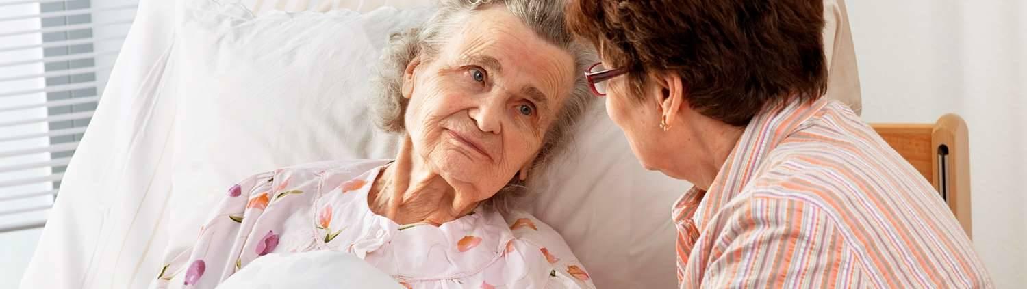 Antidekubitushilfsmittel: Alte Frau liegt im Bett und schaut einer Frau in die Augen, die neben ihr sitzt.