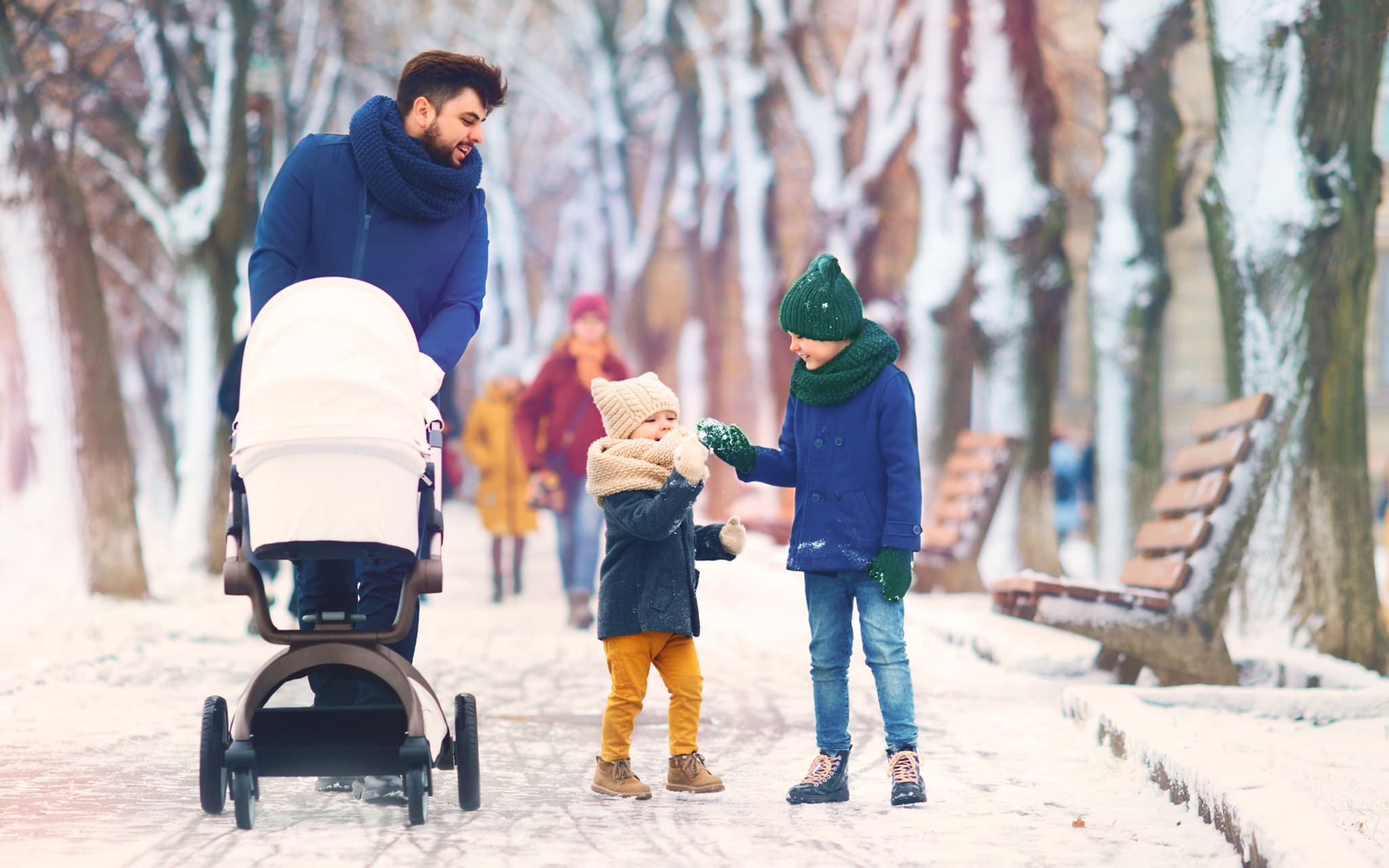 Mann schiebt Kinderwagen, zwei kleine Kinder begleiten ihn, sie spazieren im verschneiten Park