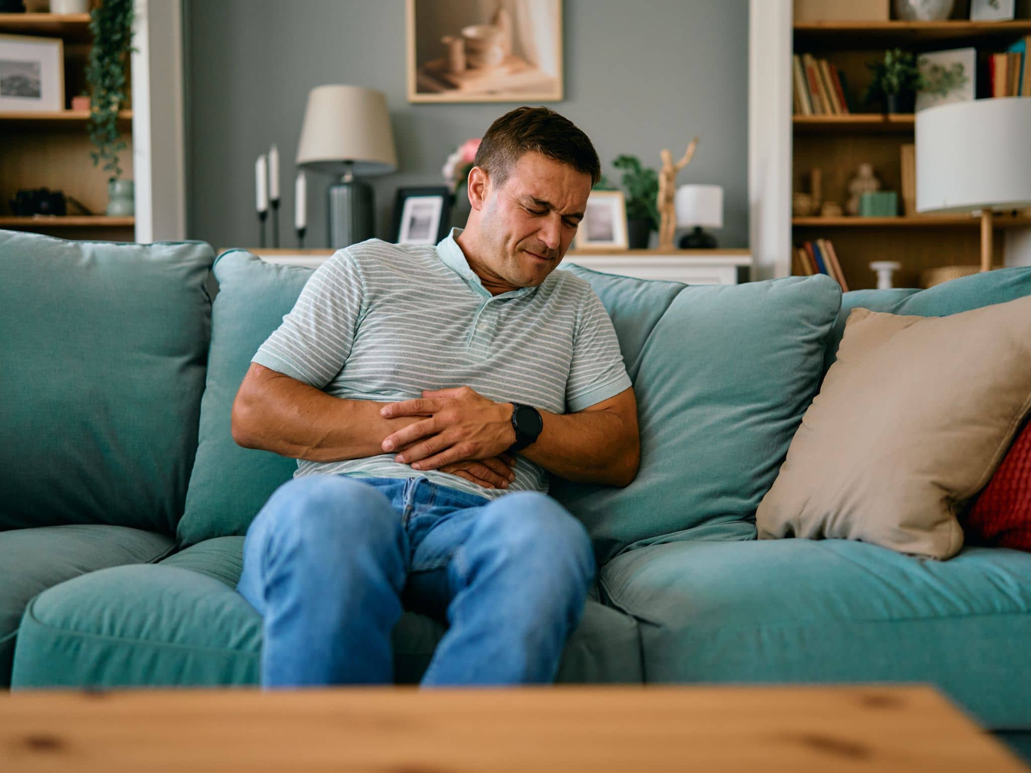 Hausmittel bei Übelkeit: Ein Mann sitzt auf dem Sofa und hält sich Hand auf den Bauch