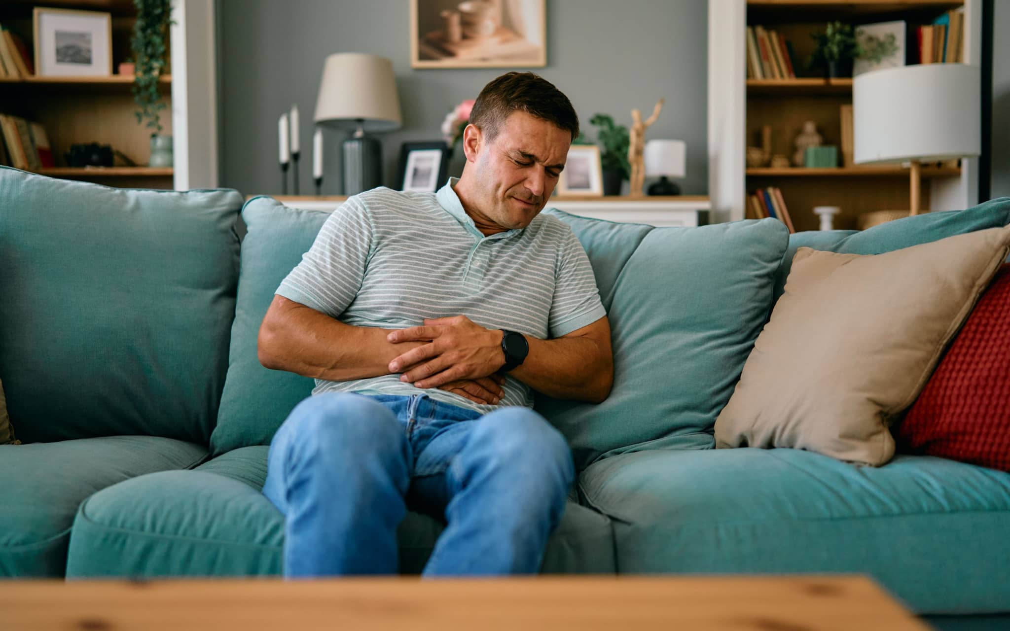 Hausmittel bei Übelkeit: Ein Mann sitzt auf dem Sofa und hält sich Hand auf den Bauch
