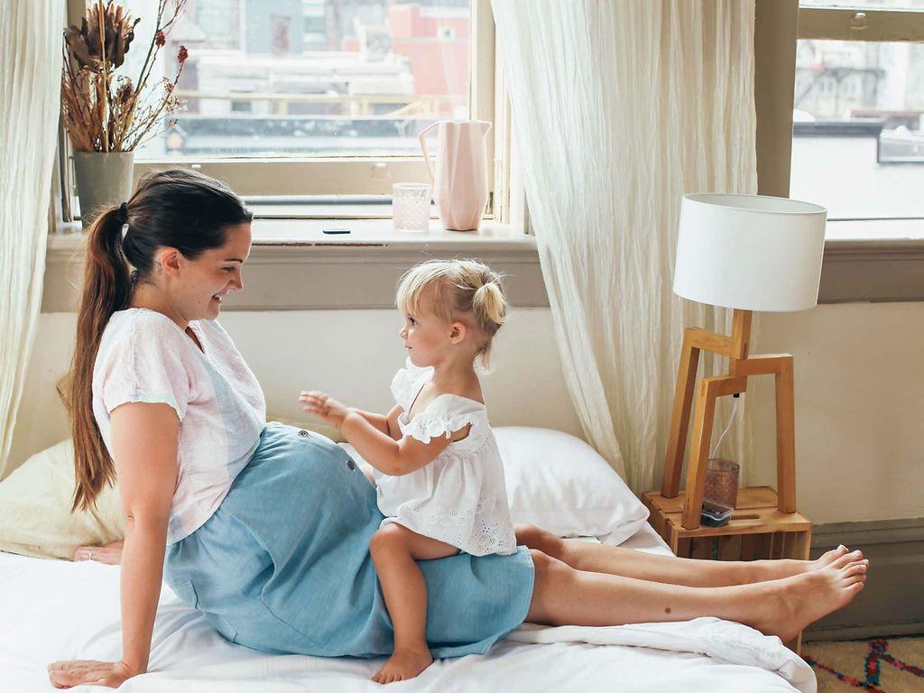 Windpocken Schwangerschaft: Junge Schwangere sitzt auf einem Bett mit einem kleinen Mädchen auf dem Schoß.