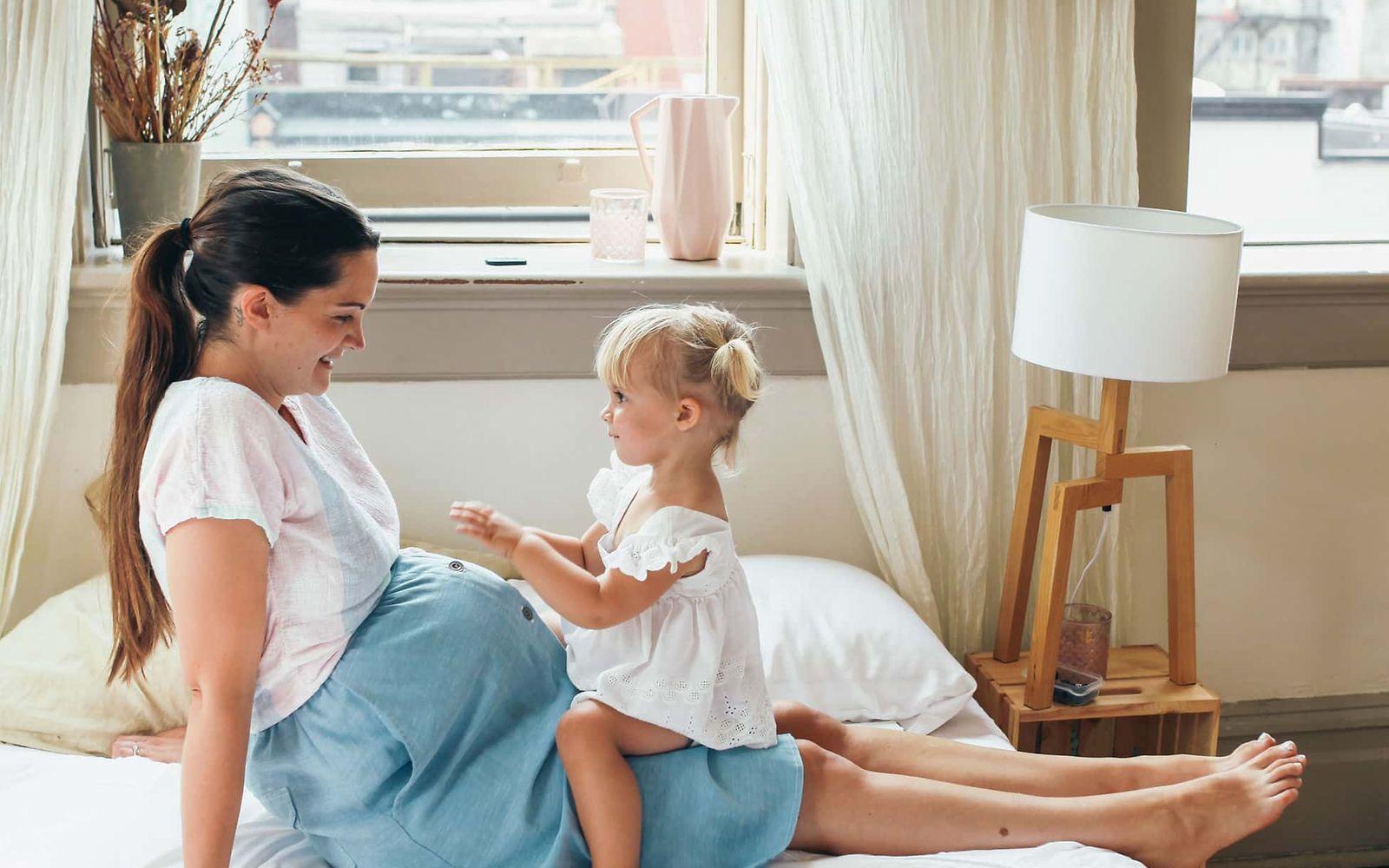 Windpocken Schwangerschaft: Junge Schwangere sitzt auf einem Bett mit einem kleinen Mädchen auf dem Schoß.