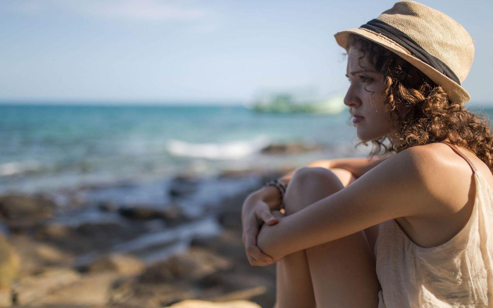 Sonnenallergie: Frau mit Hut und ernstem Gesichtsausdruck sitzt am Wasser.