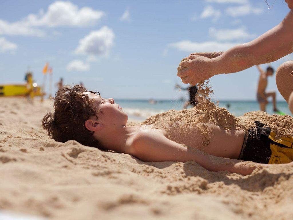 Sonnenbrand: Junge liegt am Strand in Badeshorts und wird mit Sand bestreut.