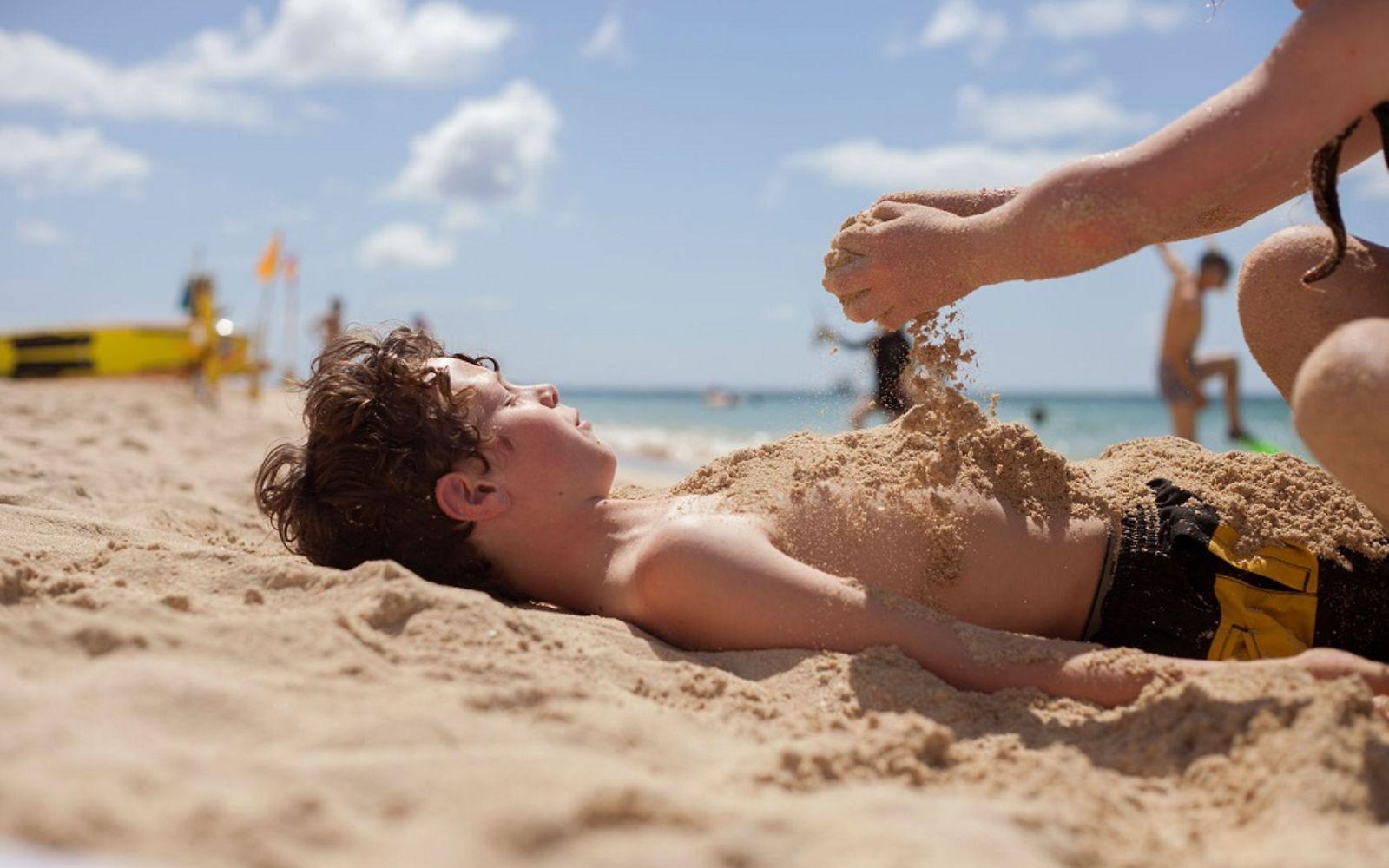 Sonnenbrand: Junge liegt am Strand in Badeshorts und wird mit Sand bestreut.