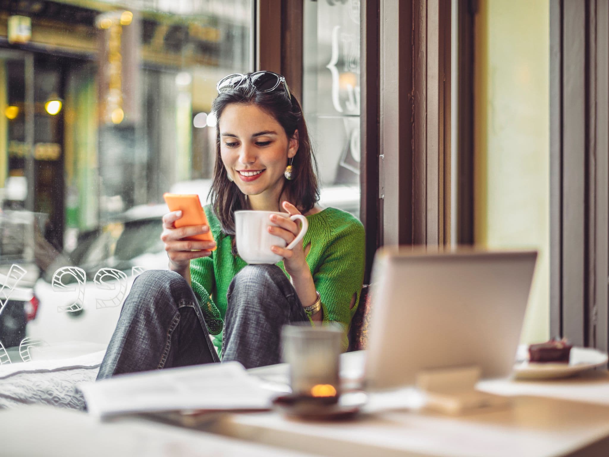 Frau sitzt im Café, guckt lachend in ihr Smartphone und trinkt aus einer Tasse.